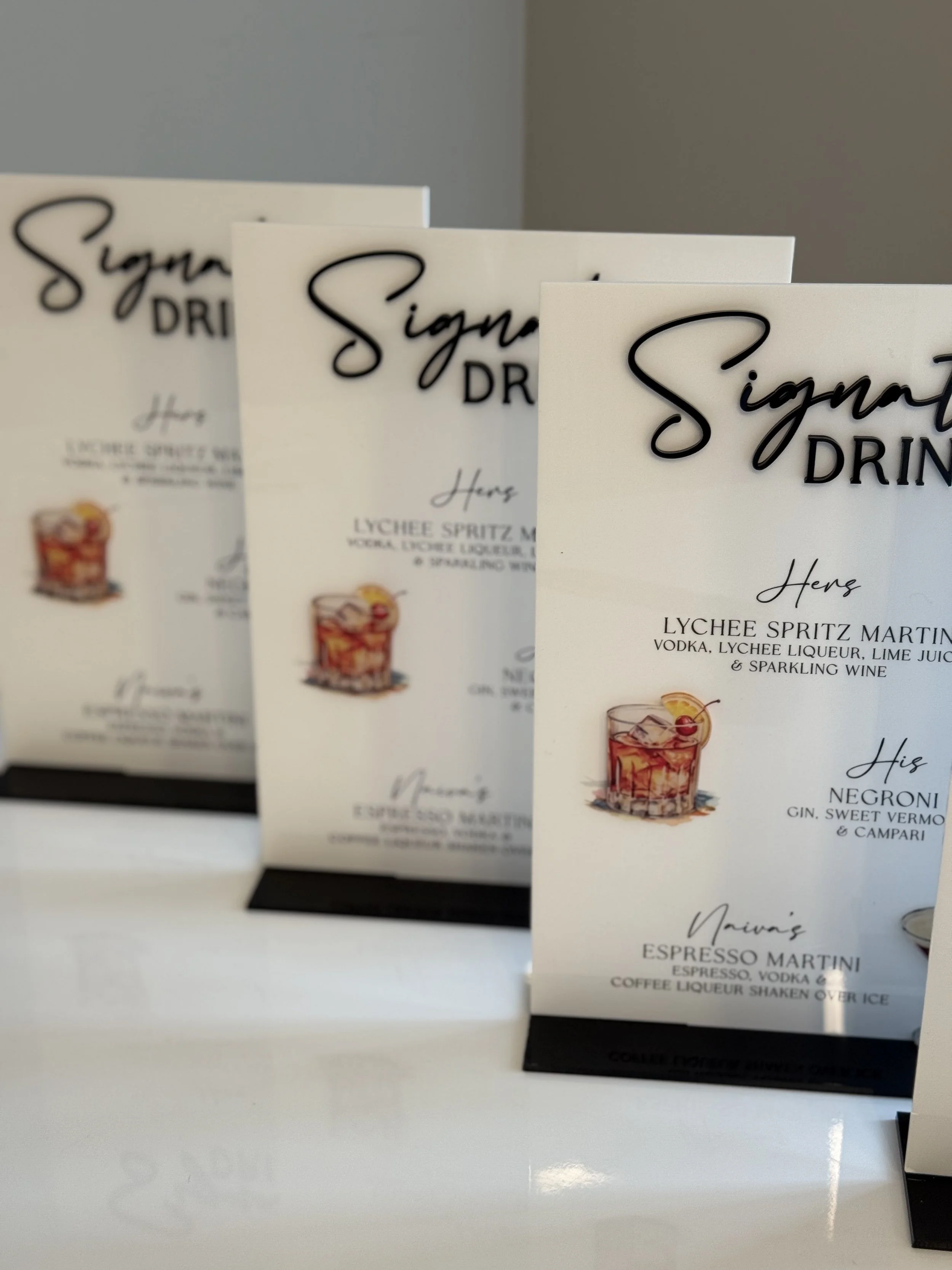 Three sign menus on a white counter displaying drink options, including lychee spritz martini, negroni, and espresso martini, with small illustrations of cocktails.