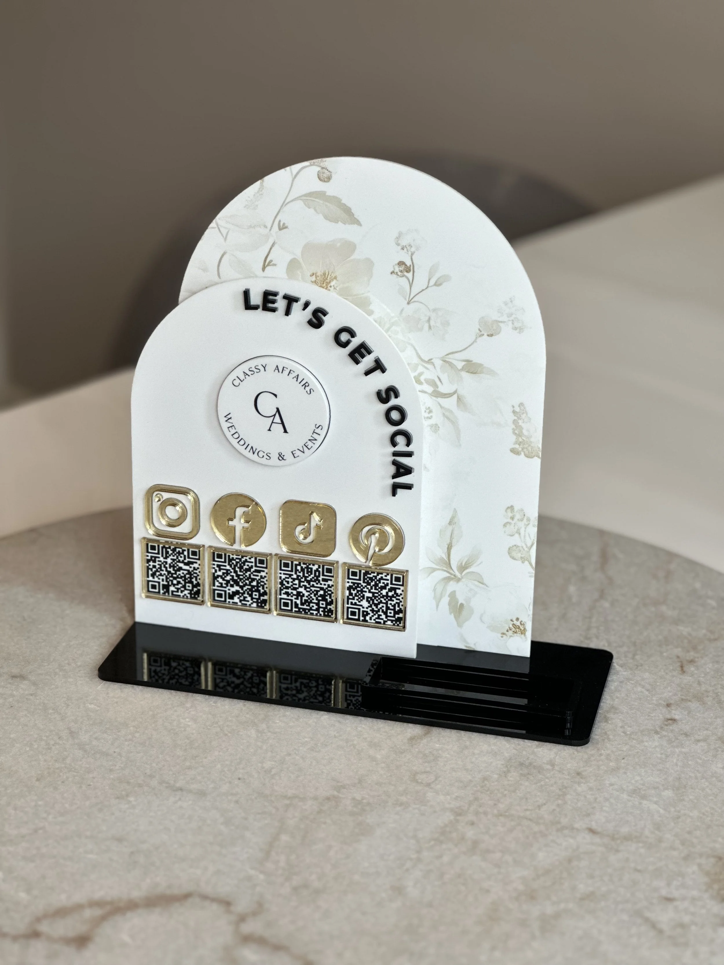 Decorative social media contact stand with icons, QR codes, and the phrase 'Let's Get Social', featuring social media logos for Instagram, Facebook, TikTok, and Pinterest, on a table surface.