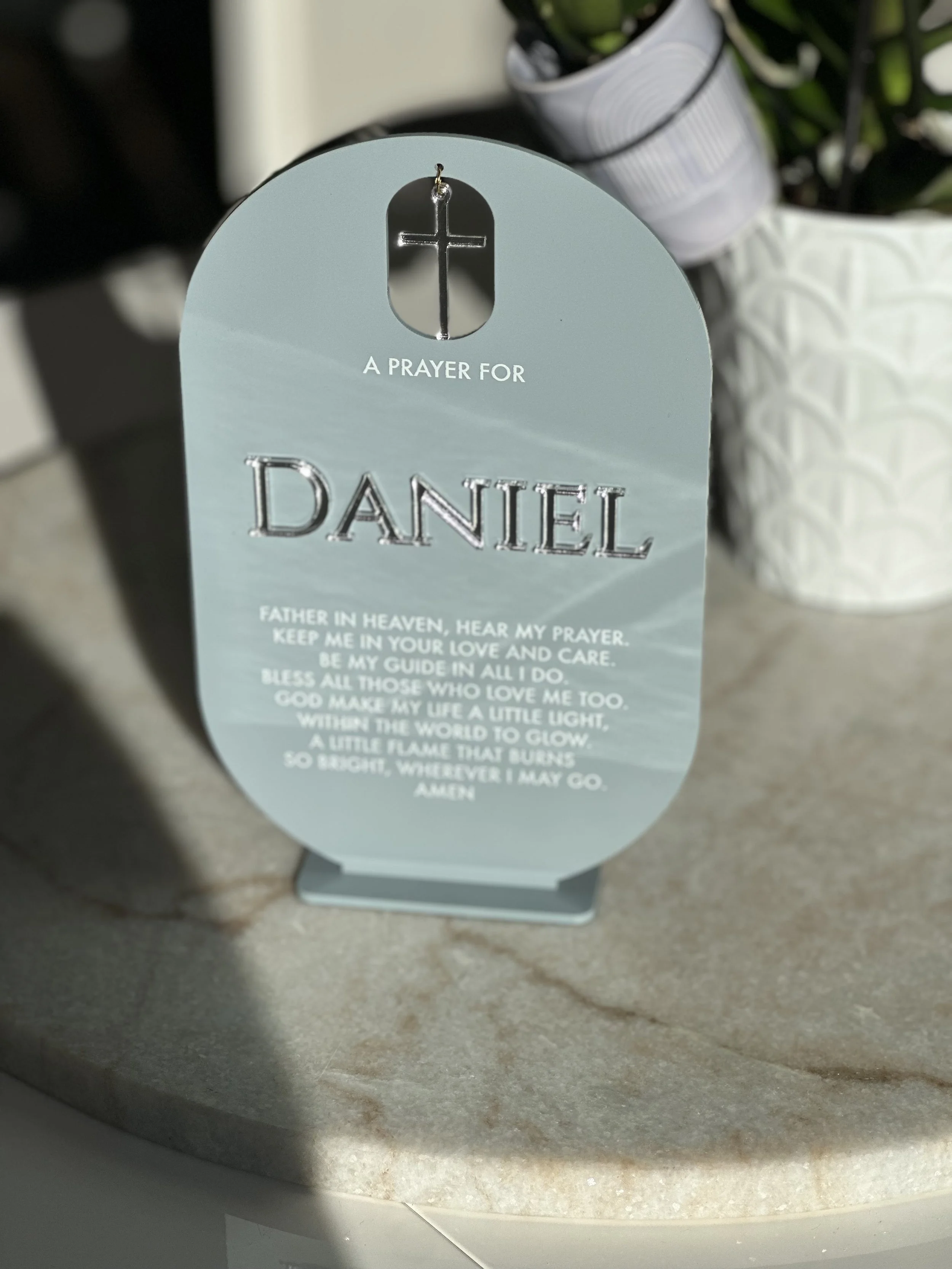 A decorative prayer card for Daniel with a cross at the top, placed on a surface with a plant in the background.