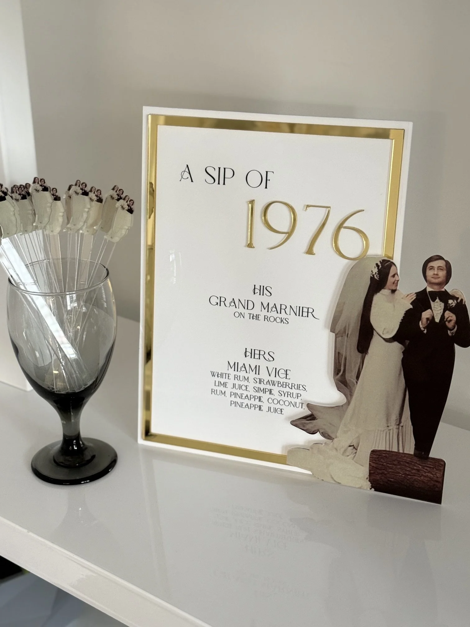A framed sign for a wedding reception with the headline 'A Sip of 1976' and a bride and groom figurine at the bottom. To the left, there is a glass vase with white flowers on toothpicks.
