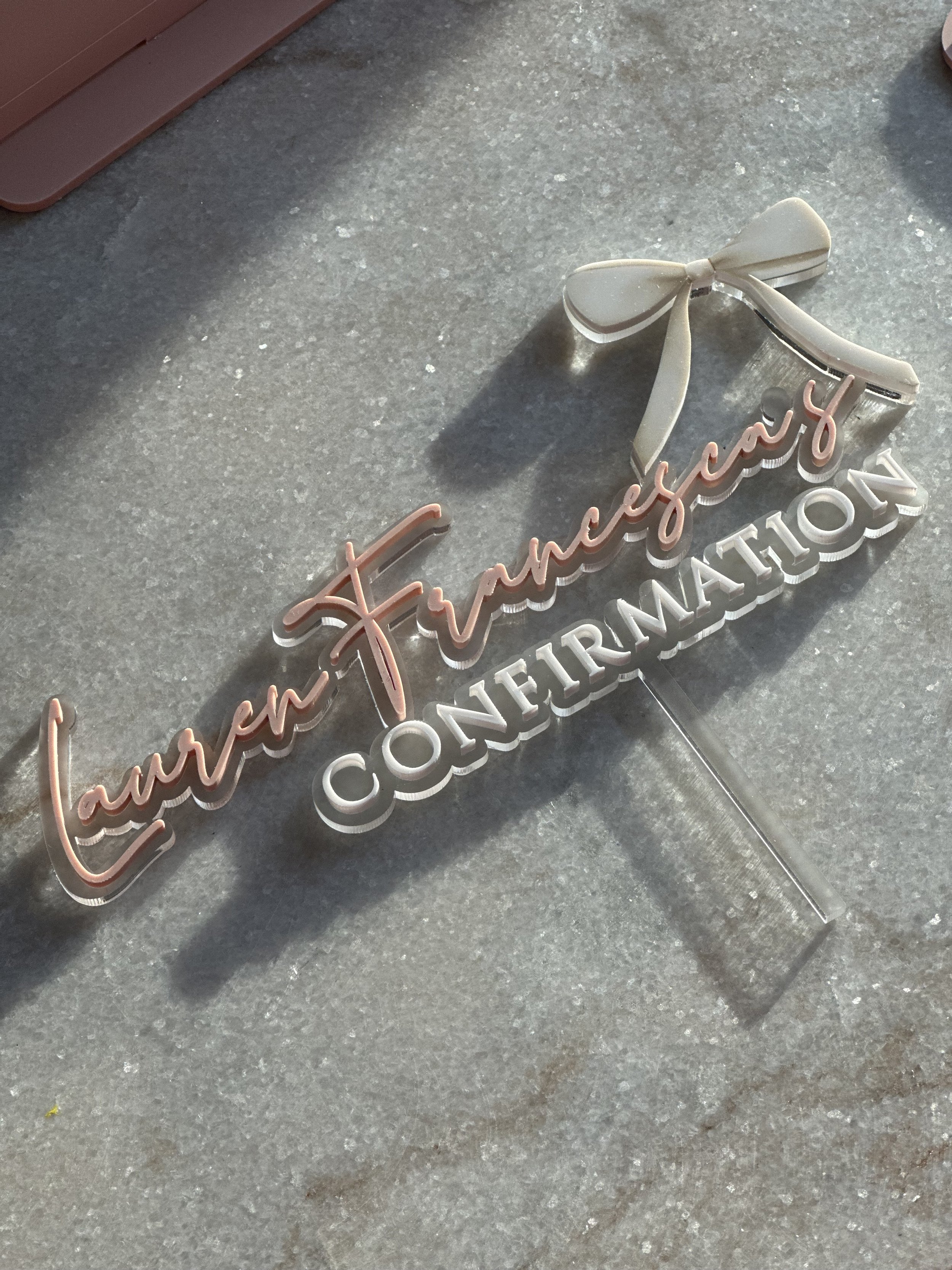 A decorative sign with the words "Lauren Francesca CONFIRMATION" in pink cursive and white block letters, with a small white bow on top, placed on a gray surface.