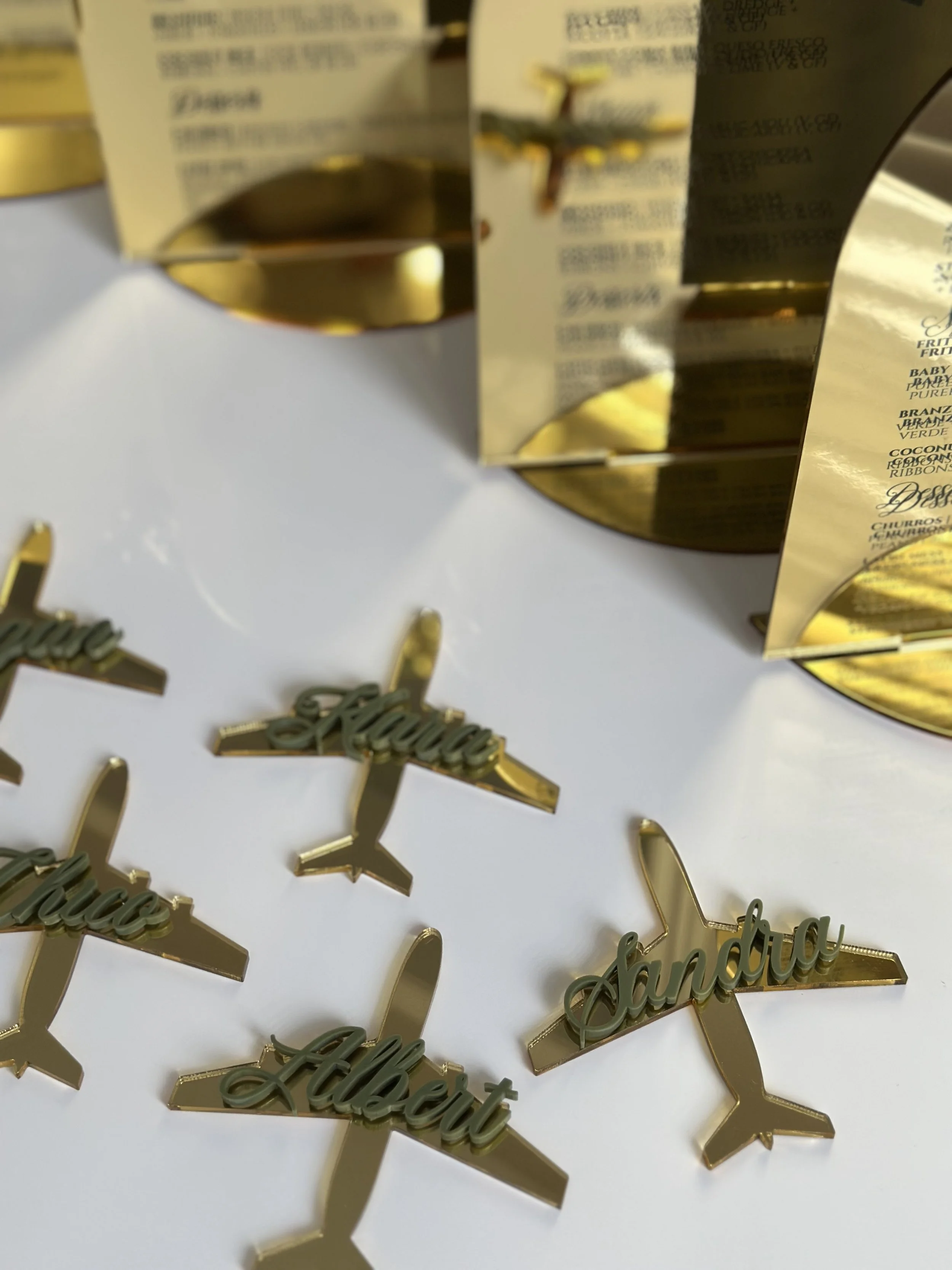 Gold airplane-shaped decorations with (from left to right) names "Aubrey", "Albert", and "Sandra" in cursive script placed on a white surface, with blurred boxes in the background.