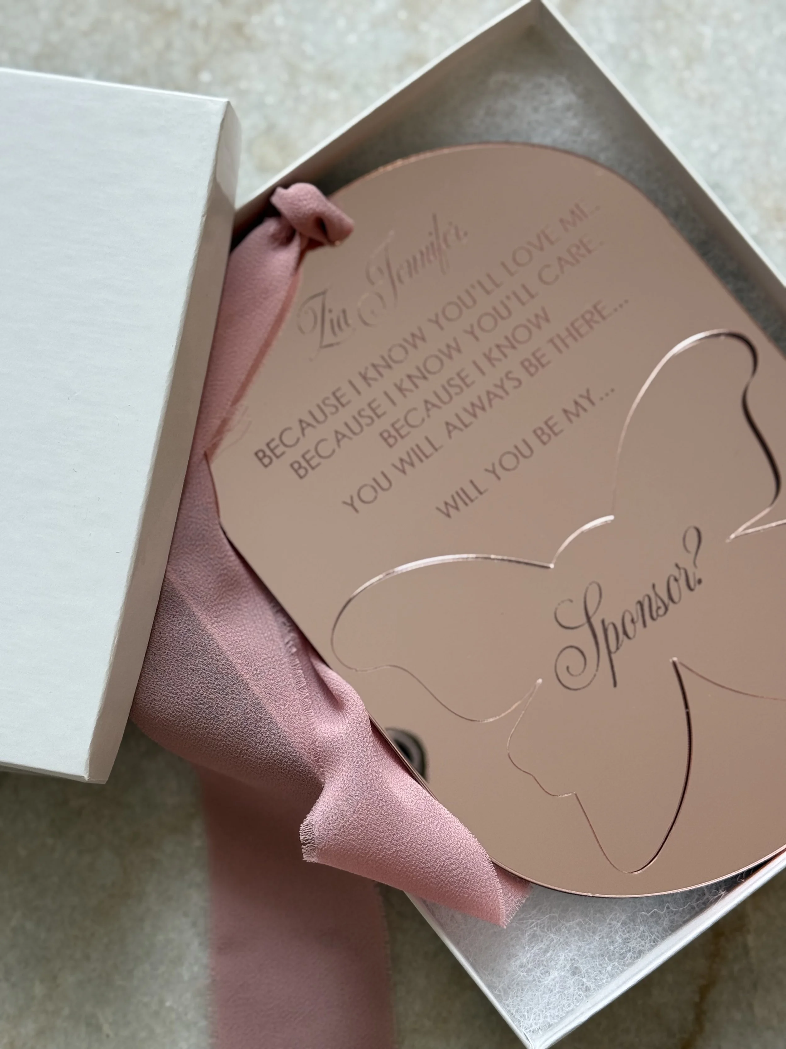 A pink card inside a white gift box, with a pink ribbon, featuring a heartfelt message and a space for a sponsor's name.