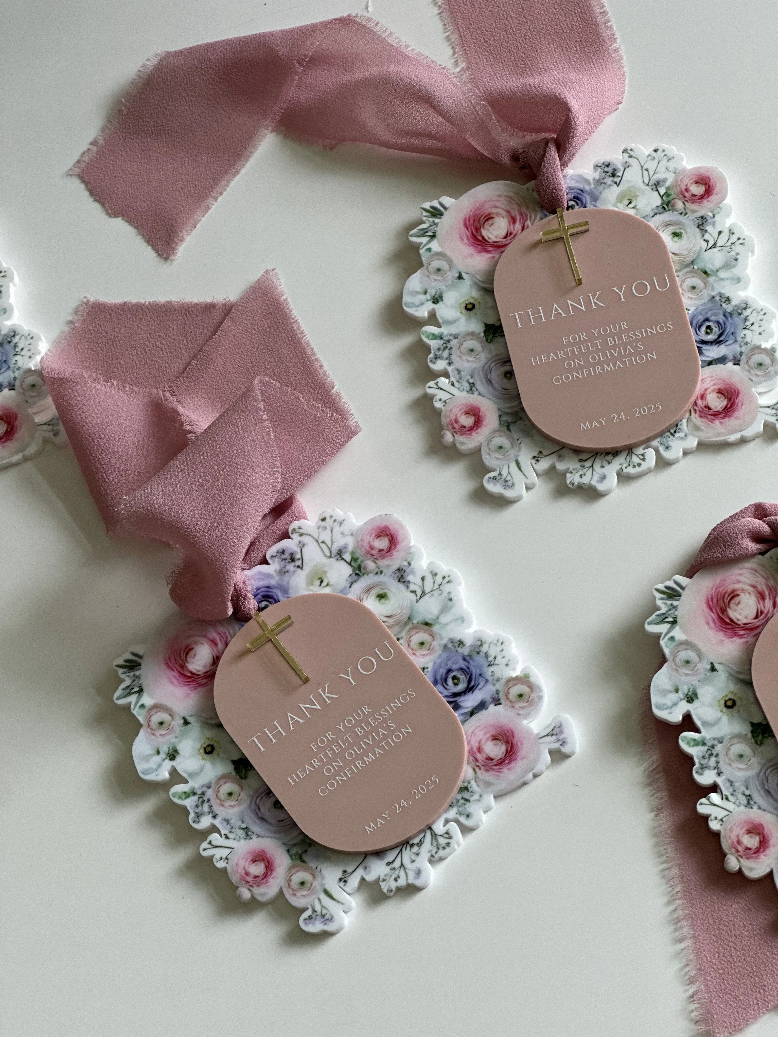 Three floral thank you tags with pink ribbons and a small gold cross, surrounded by paper cutouts of flowers in pink, purple, and white, on a white surface.