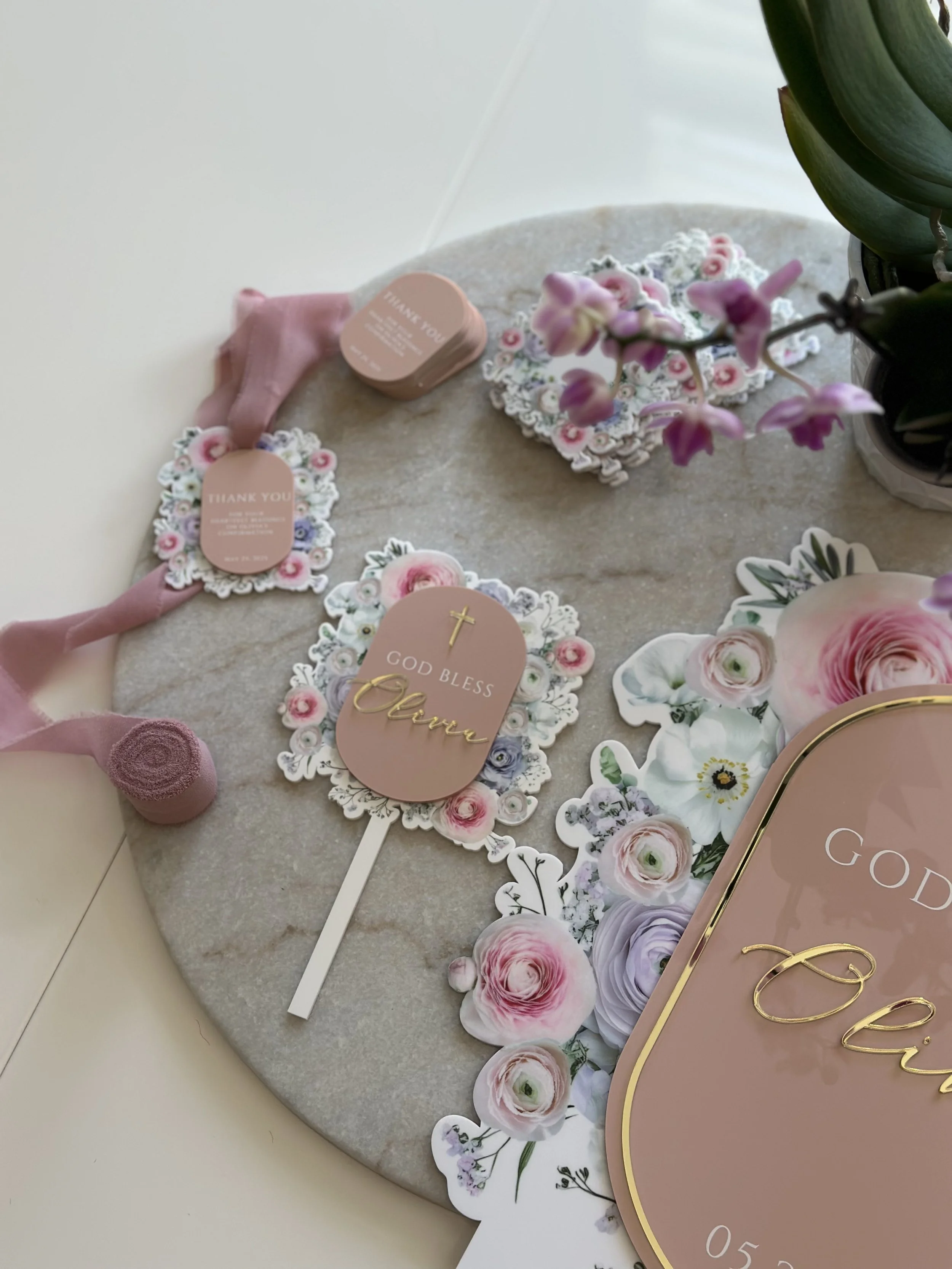 A round stone display with floral stickers, pink thank you tags, a pink 'God Bless Mom' sign with a cross and gold lettering, pink ribbon, and a pink flower sticker, along with a pink potted plant with purple flowers.