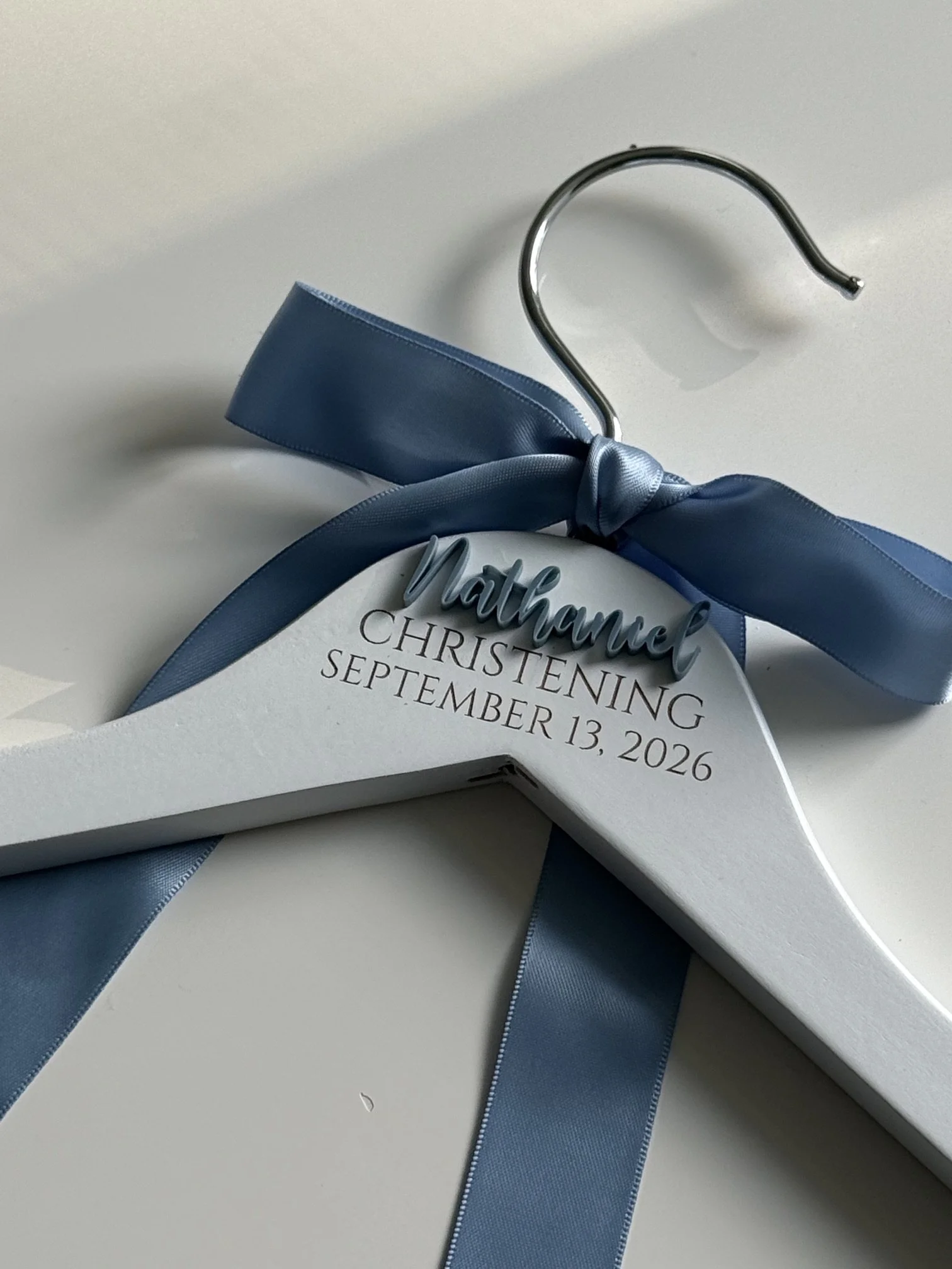 A white heart-shaped card with a blue satin ribbon tied in a bow around it, displaying the name 'Matthew' and details of a christening event scheduled for September 13, 2026.