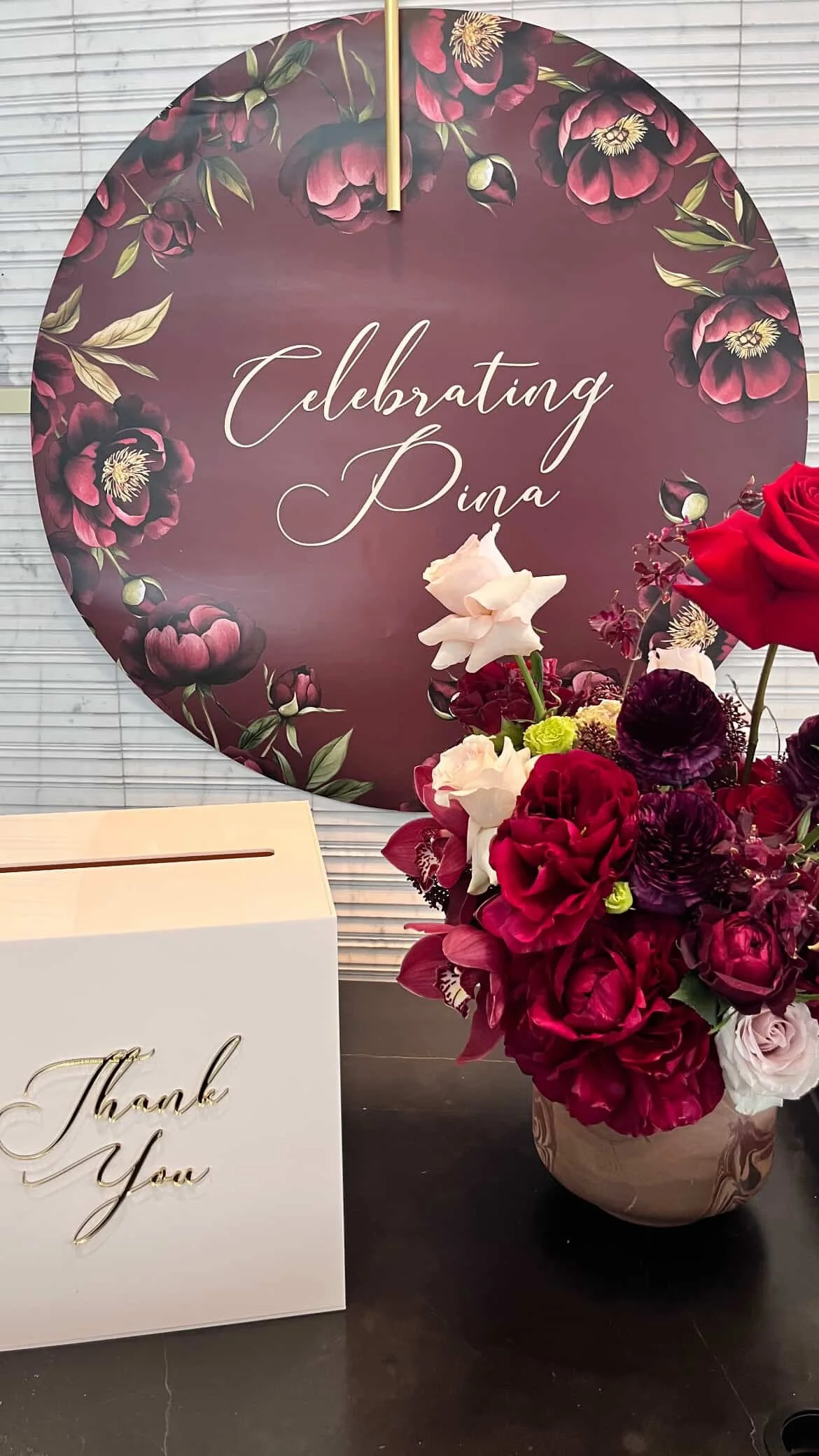 A floral arrangement with red, pink, and purple flowers in a marble-patterned vase, placed next to a white 'Thank You' card with gold cursive letters, and behind them is a round sign with a floral border and the words 'Celebrating Dina' in cursive.
