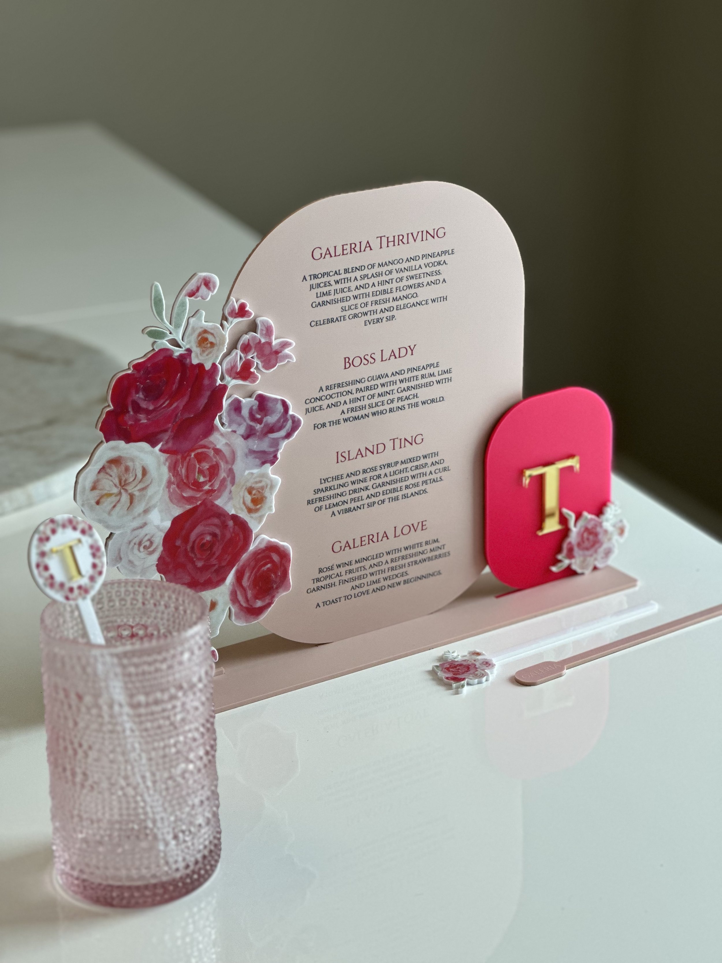 Pink floral-themed drink menu displayed on a table with decorative elements, including a pink monogram letter 'T' and floral accents.