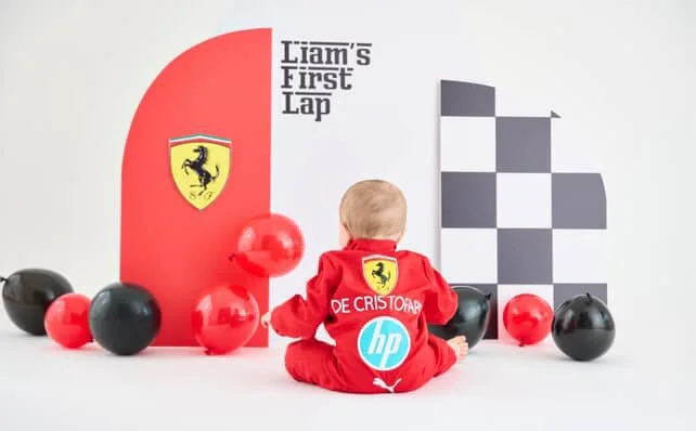 Young child dressed as a race car driver sitting on the floor with multiple red and black balloons, facing a wall with a sign that says 'Liam's First Lap' and a Ferrari logo, surrounded by black and red balloons.