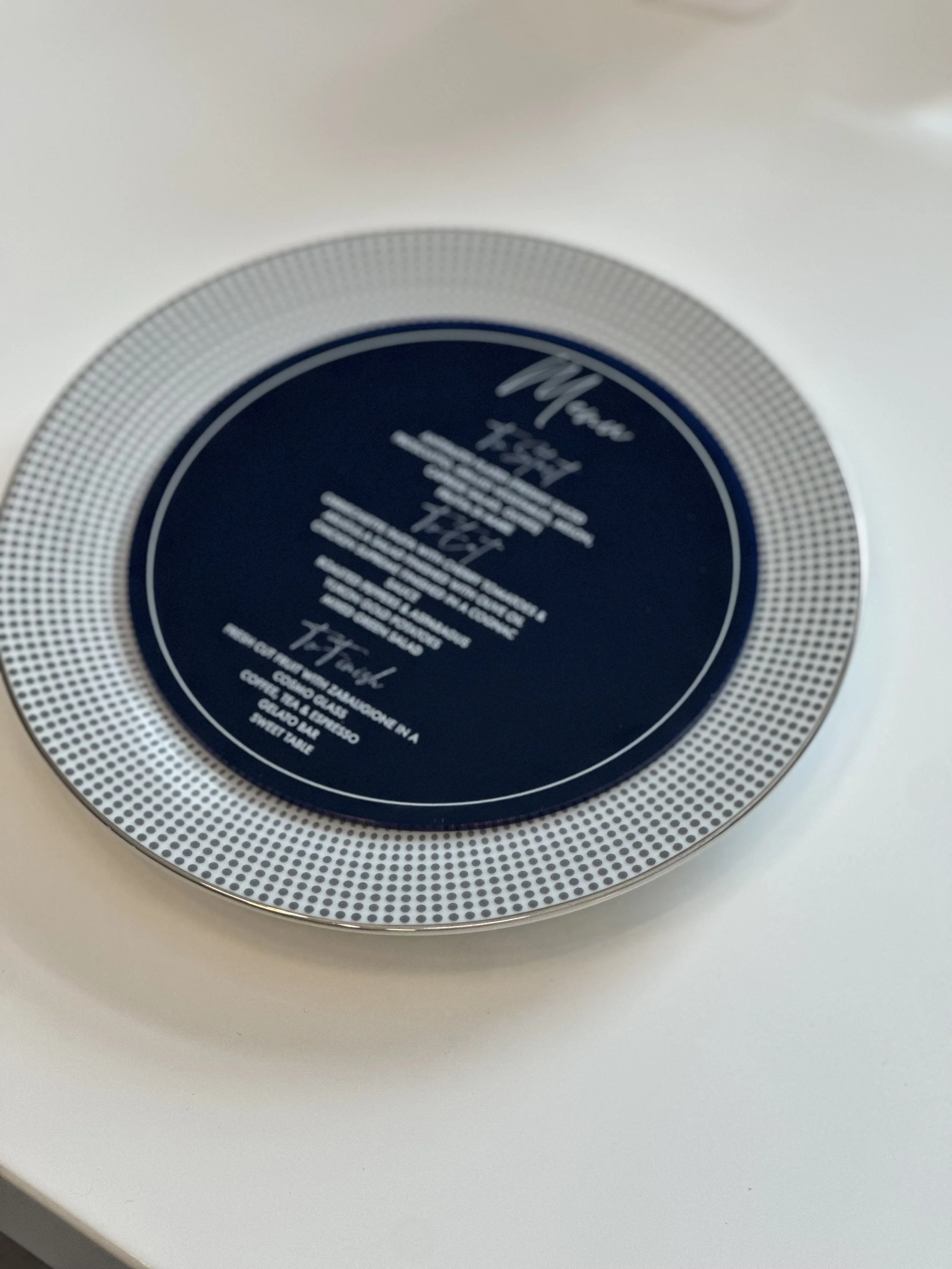 A round plate with a silver dotted border and a dark blue center, featuring white cursive and print text.