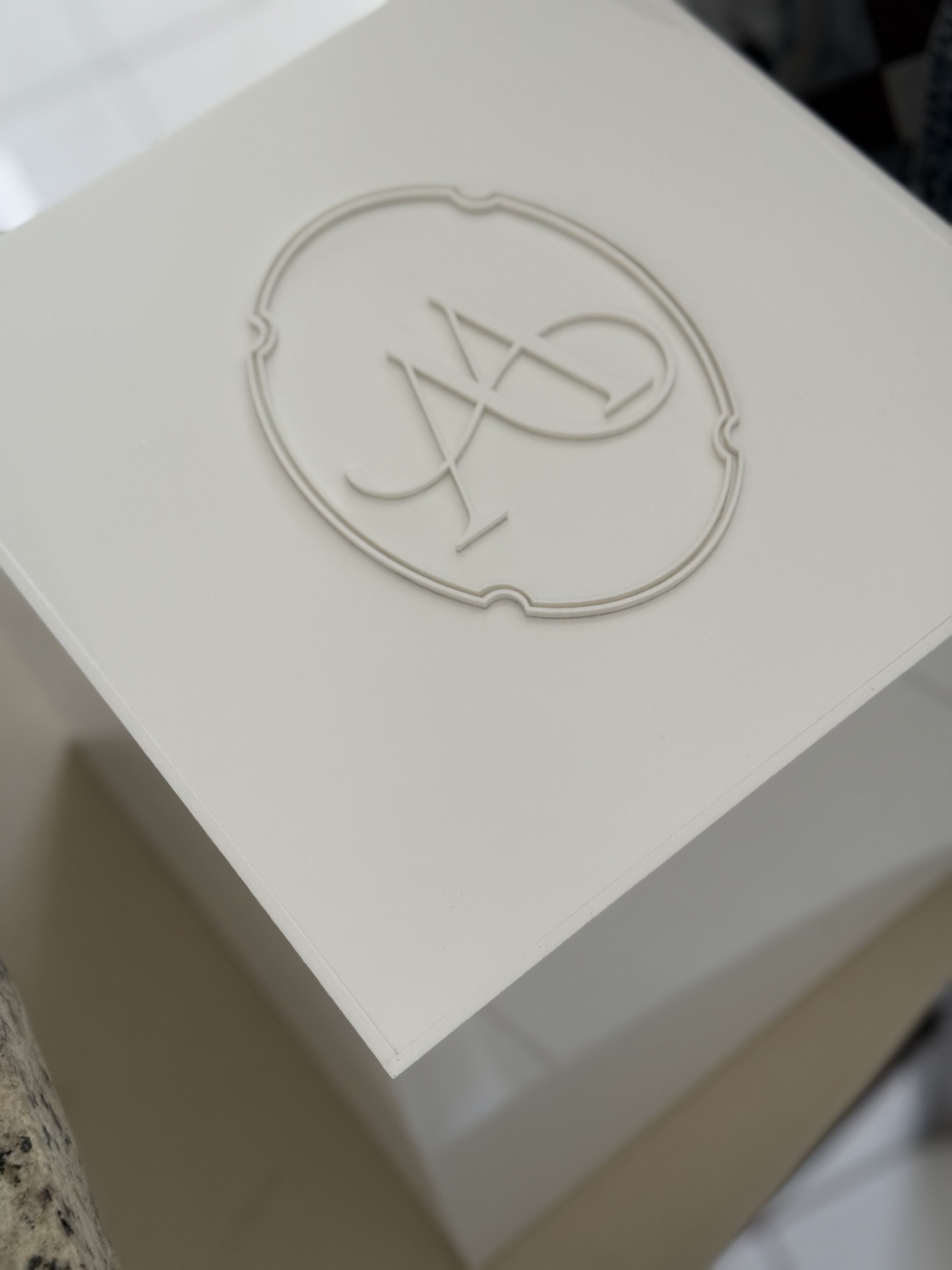 A white box with embossed logo on the lid, featuring a decorative circle with a stylized letter 'A' inside.
