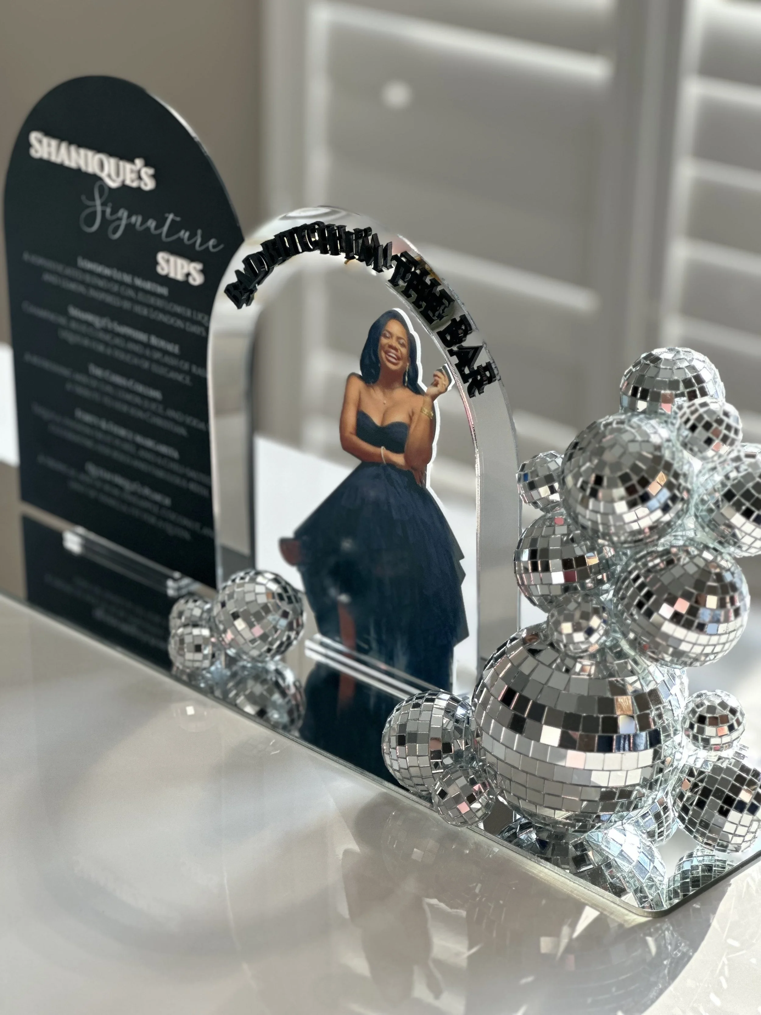 A decorative acrylic mirror with a cutout photo of a woman in a black dress, surrounded by silver disco ball ornaments. The mirror has a black sign on the left with text about a signature drink, and a black arch with the words "You Are the Bar" at th
