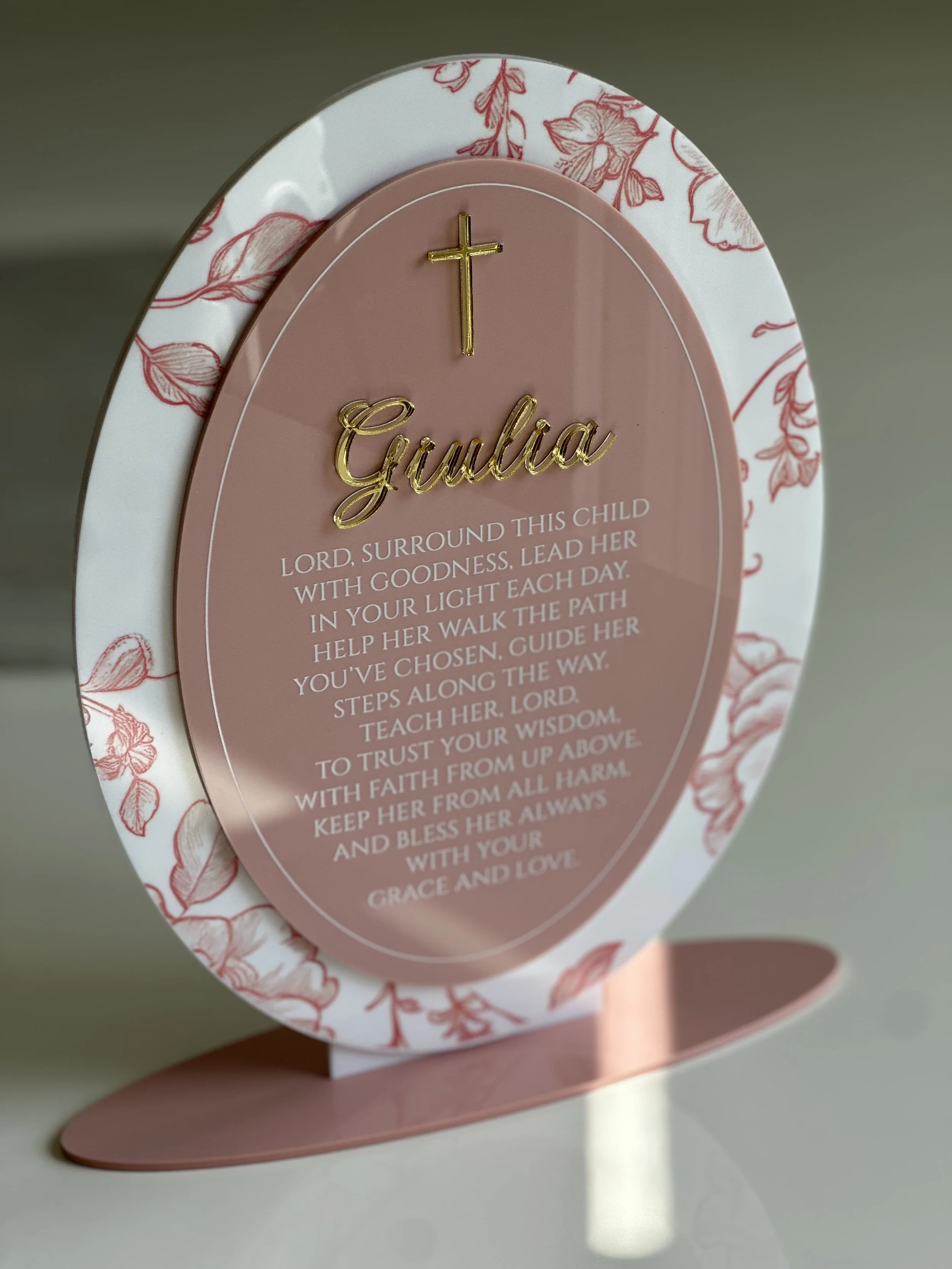 Decorative standing plaque with a pink background, gold cross and name 'Gulia', and a prayer for a child's protection and guidance. The plaque has a white border with red floral designs.
