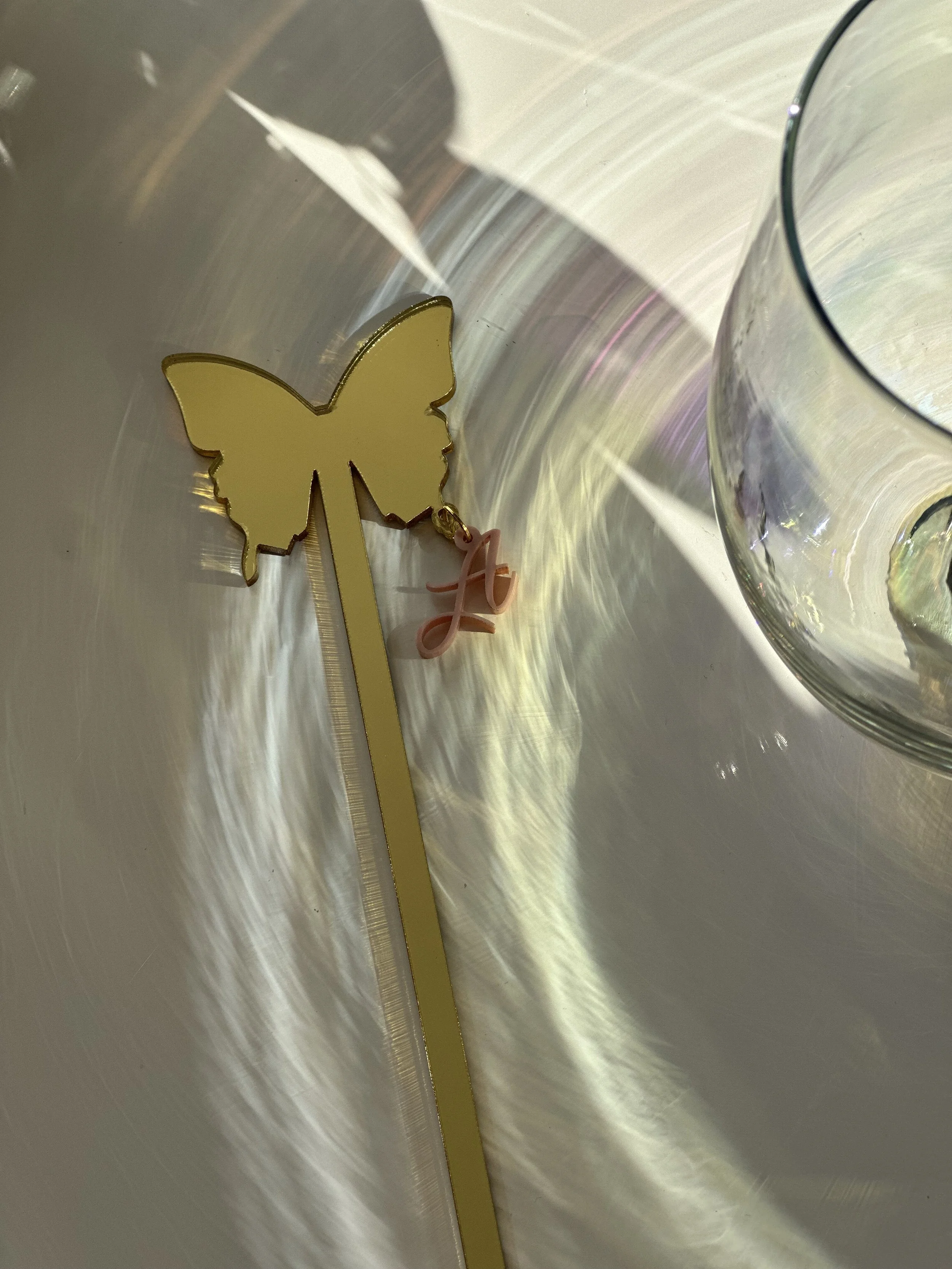 Yellow butterfly-shaped decoration with a pink letter 'L' charm, placed on a reflective surface next to a glass container.