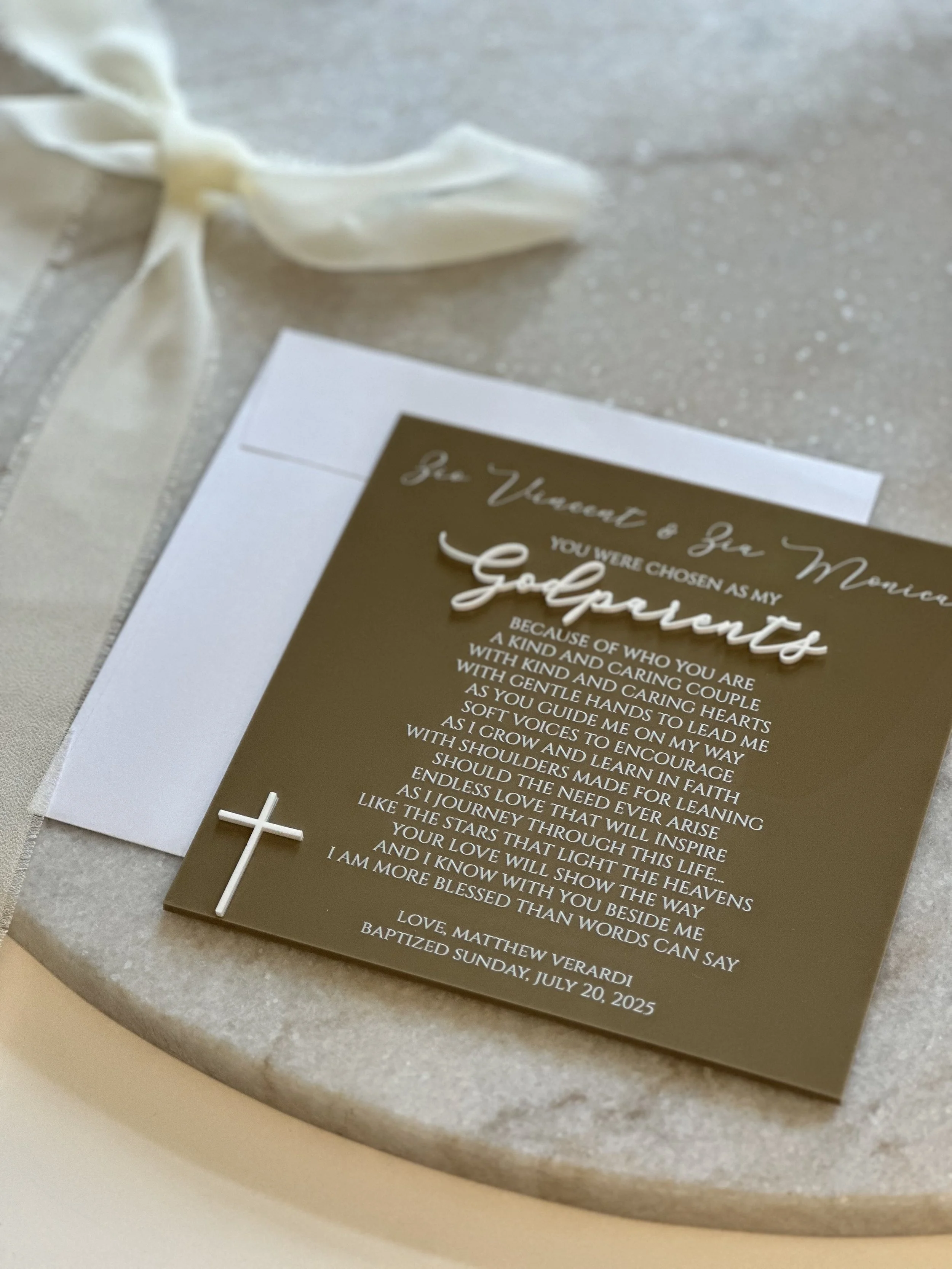 A baptism invitation card with a small white cross on the lower left corner and a white ribbon attached. The card has gold and white text welcoming guests to a baptism ceremony.