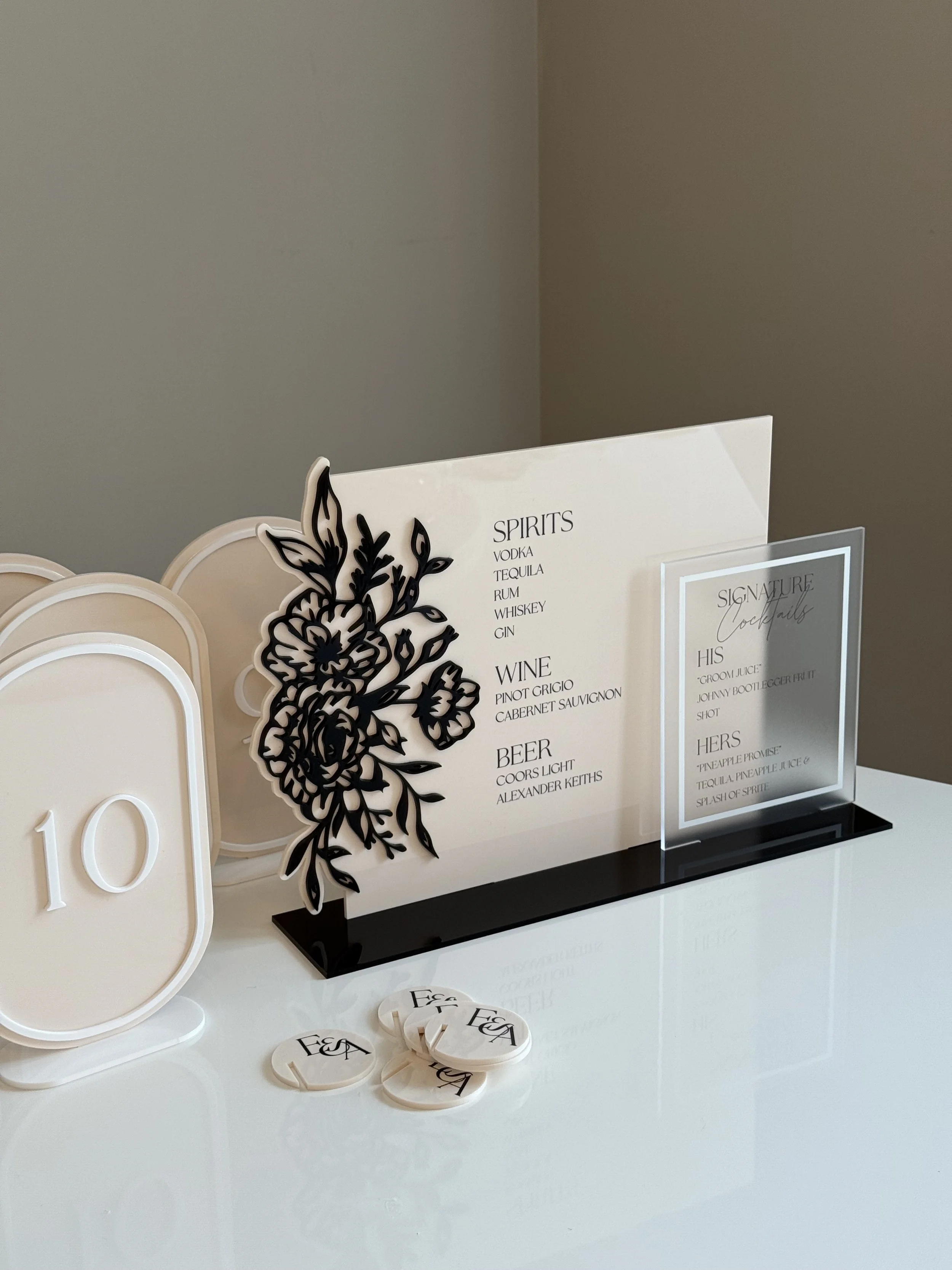 Table sign with a black floral design and drink menu listing spirits, wine, and beer, along with a smaller clear sign with cocktail names and a white table number sign showing the number 10.