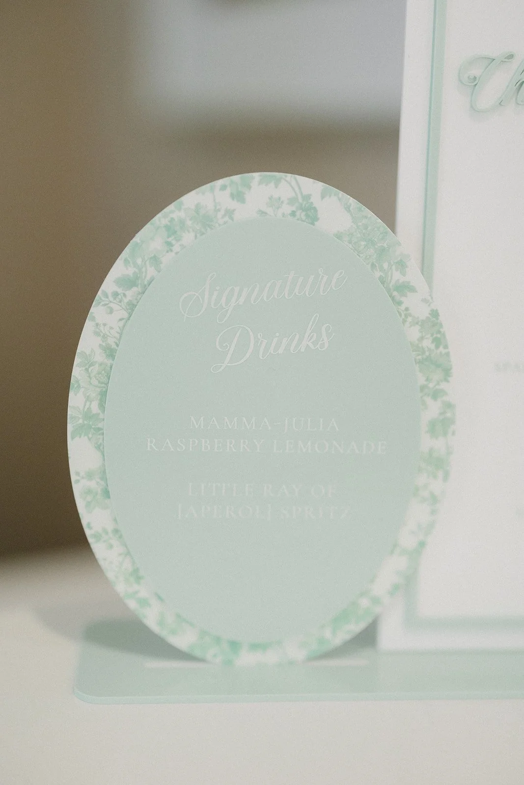 A sign with a light green floral border and white cursive text listing signature drinks, including Mamma Julia Raspberry Lemonade and Little Ray of Laporous Spritz.