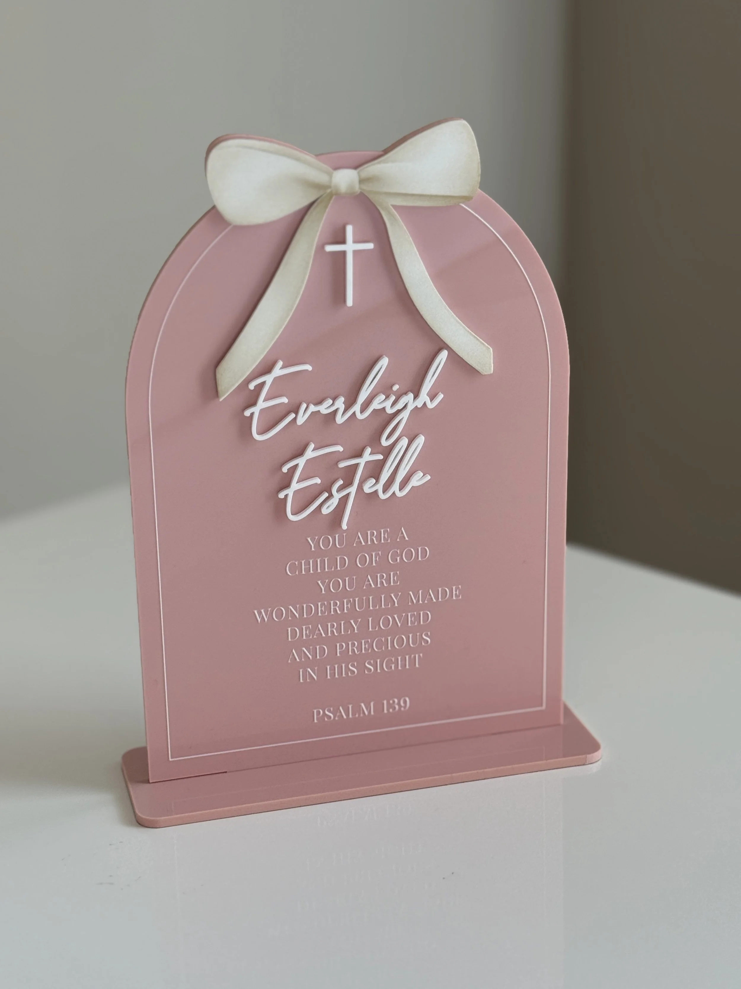 Pink decorative plaque with a white cross and a cream-colored bow at the top. The plaque has the name "Everleigh Estelle" and a Bible verse from Psalm 139, written in white and pink text.