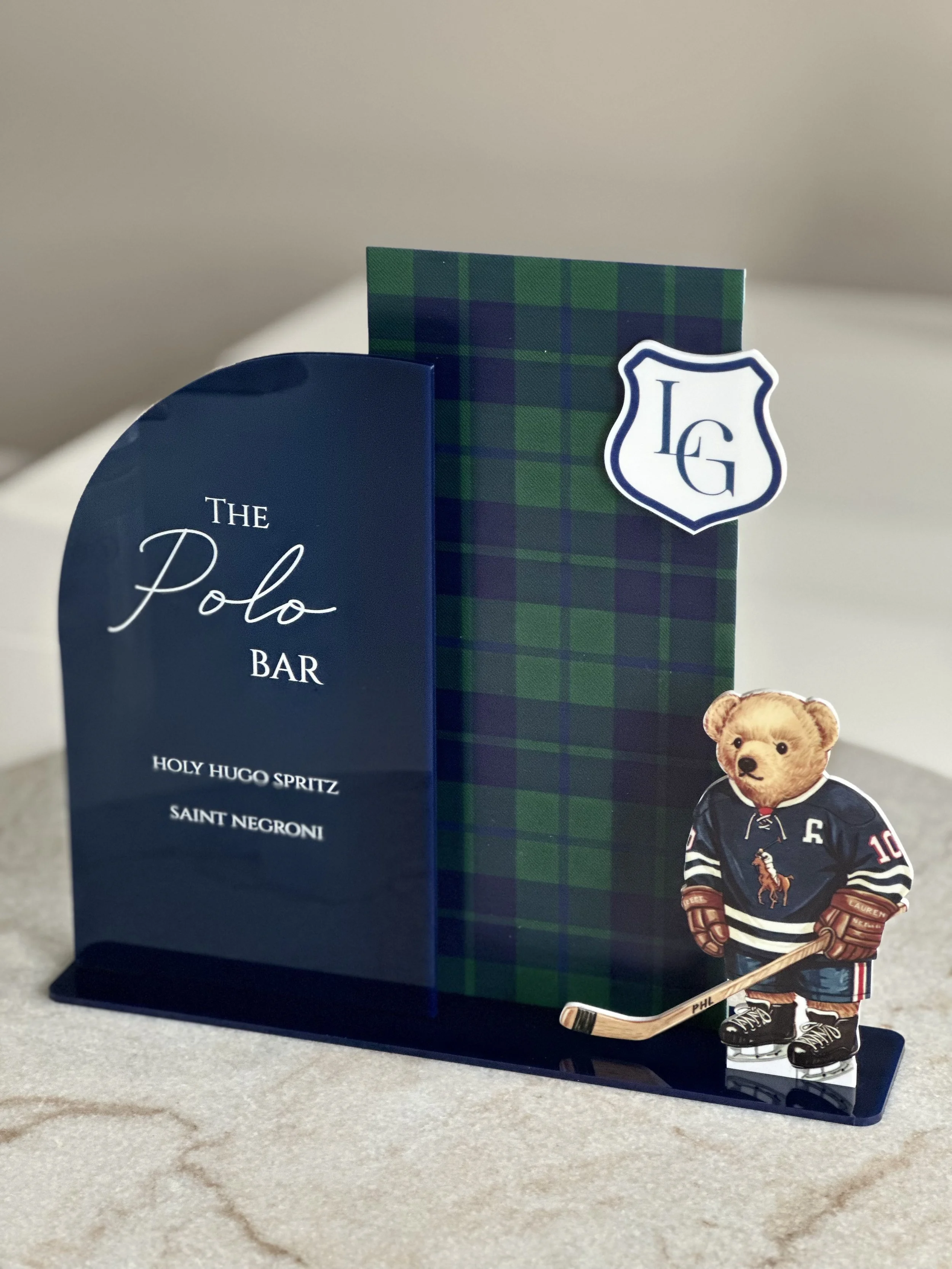 Decorative stand featuring a sign for 'The Polo Bar' with a menu listing 'Holy Hugo Spritz' and 'Saint Negroni', a plaid-patterned box with a crest logo, and a cute bear figurine dressed in a hockey jersey holding a hockey stick.