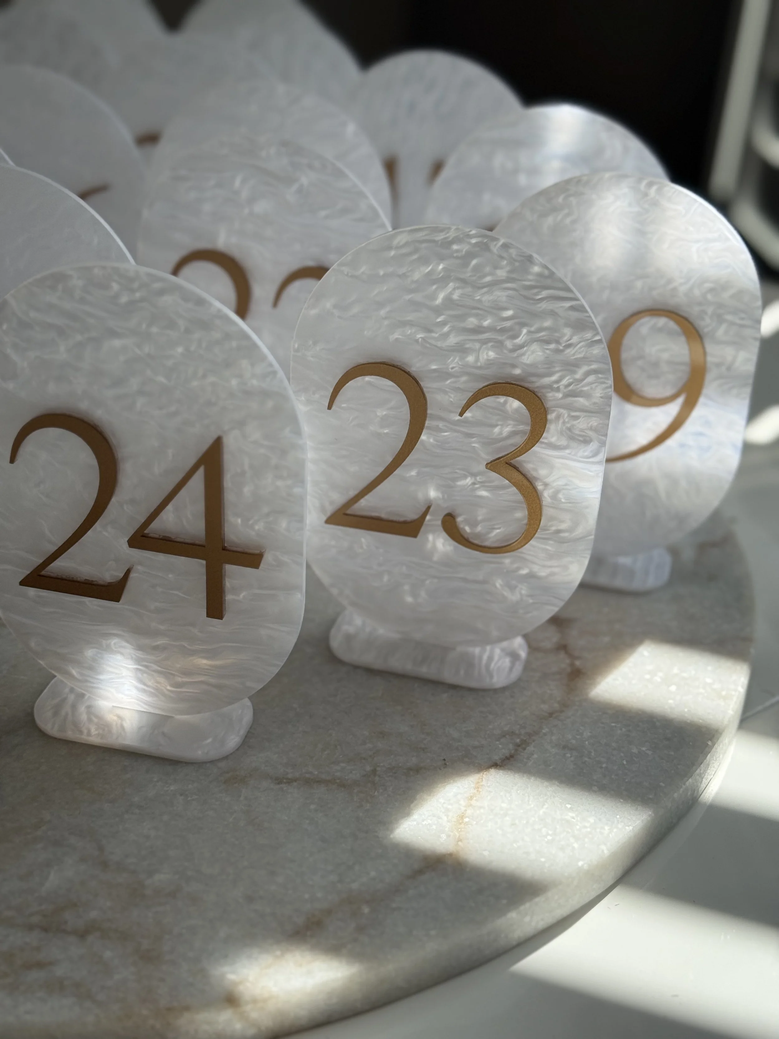 Numbered table markers with gold numbers on a white pearl-like background, arranged on a round stone surface.