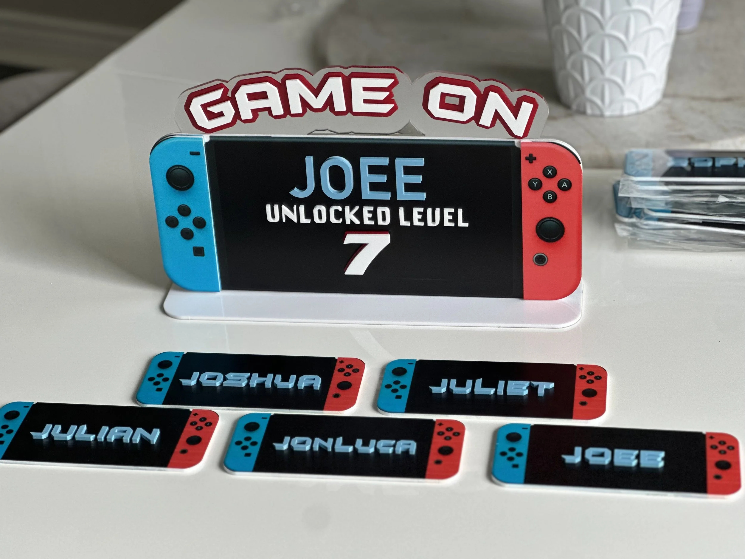 A Nintendo Switch gaming console with a display reading "JOEE UNLOCKED LEVEL 7" behind a stand featuring a "GAME ON" sign. There are custom name tags in front of the console reading "JOSHUA," "JULIAN," "JONLUCAS," "JULIET," and "JOEE."