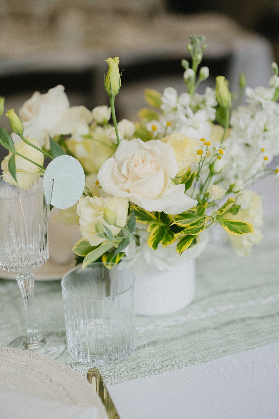A floral centerpiece with white roses, small white and yellow flowers, and green leaves, placed in a white vase on a table with glasses and a tablecloth.