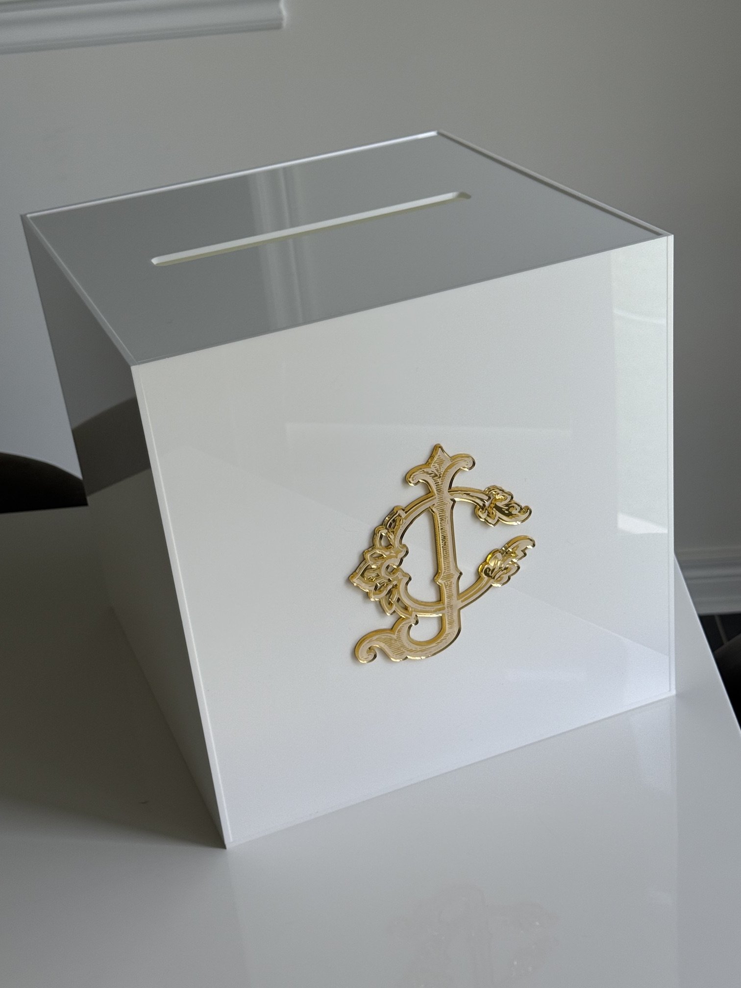 A white box with a gold decorative logo on the front, featuring a large ornate letter "C" and other decorative elements, sitting on a plain surface.
