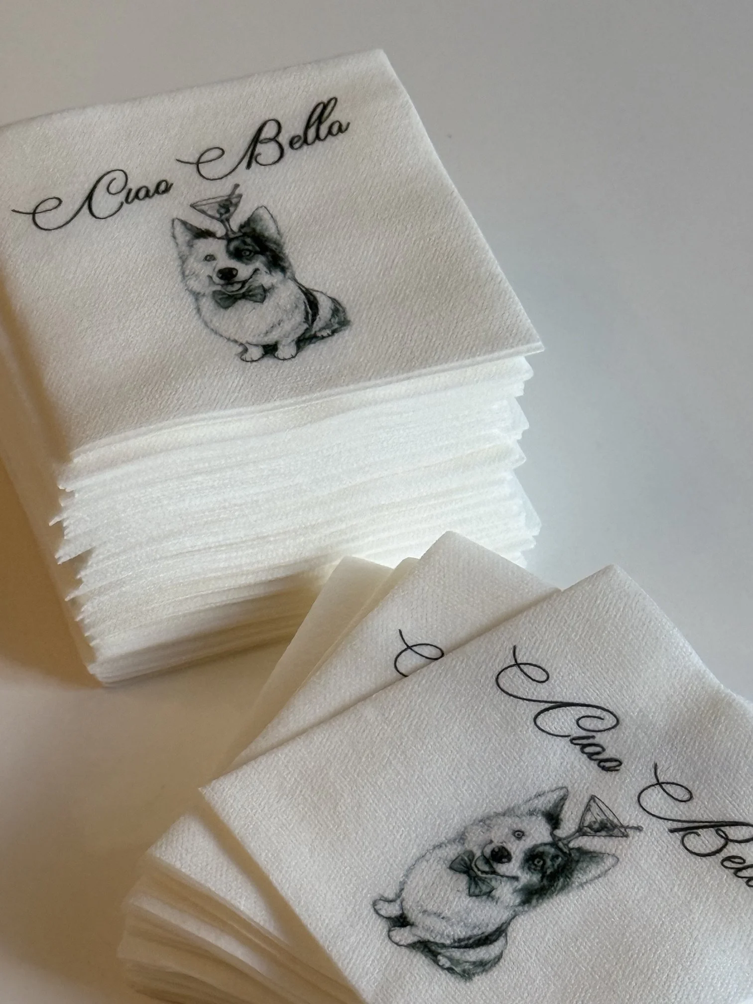 Stack of white napkins featuring a printed illustration of a dog with a butterfly on its head and the text 'Ciao Bella' in elegant cursive.