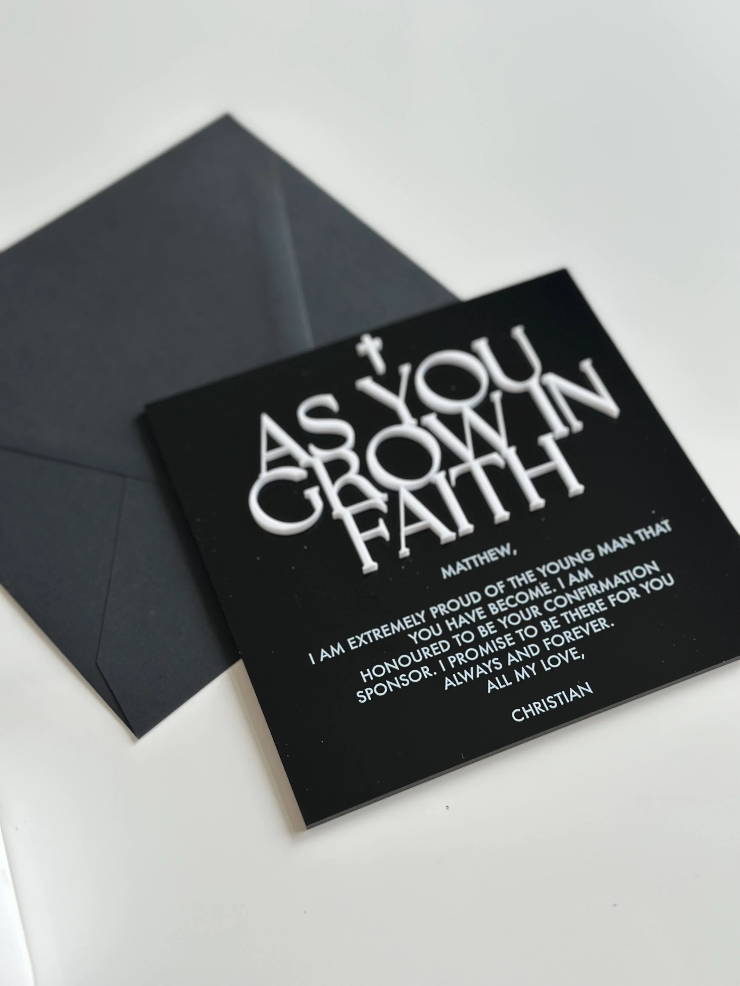 A black greeting card with white text and a small cross at the top, placed next to a black envelope, on a white surface. The card features a religious message expressing pride and love from Christian parents to their son.