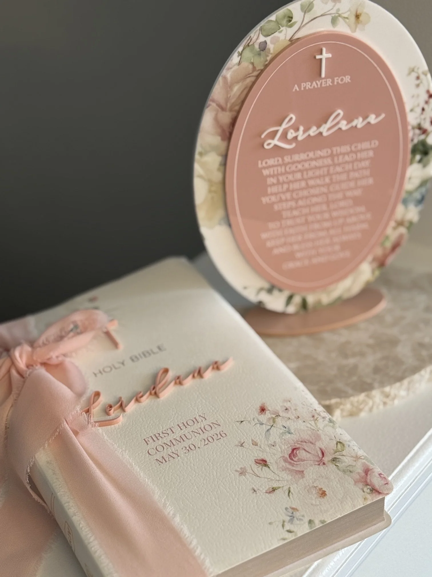 A decorative Bible with pink satin ribbon and embroidery, and a floral card with a pink circular plaque containing a prayer for a loved one, dated May 30, 2026.