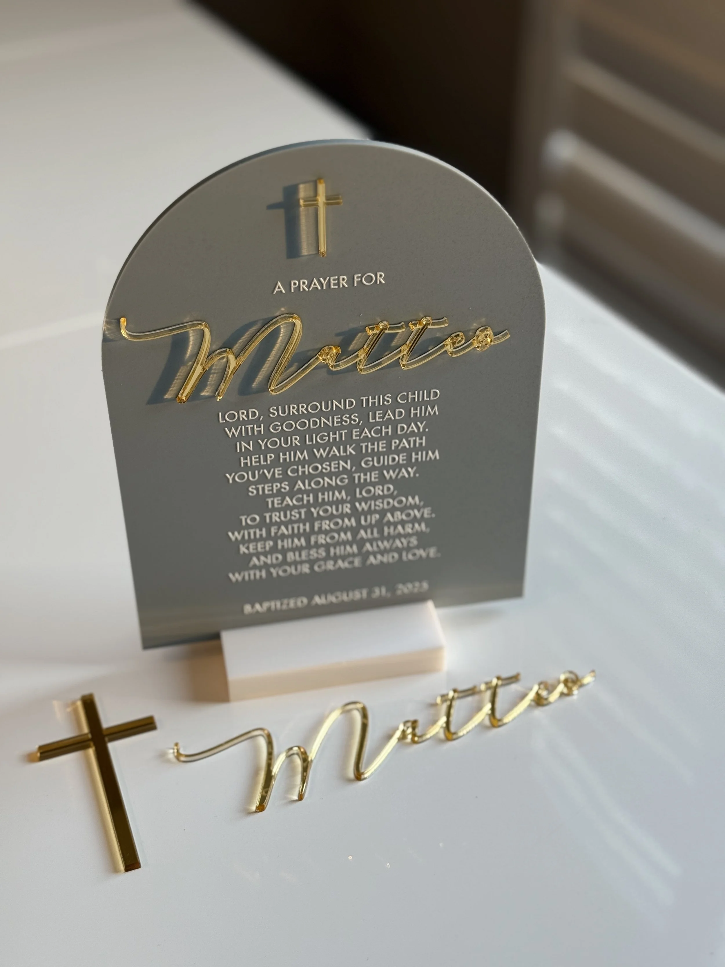 A decorative religious display with a gray plaque and gold text, mounted on a white stand. The plaque features a gold cross at the top and the word 'Matte' in gold cursive. A separate gold cross and the word 'Matte' are laid in front of the display.
