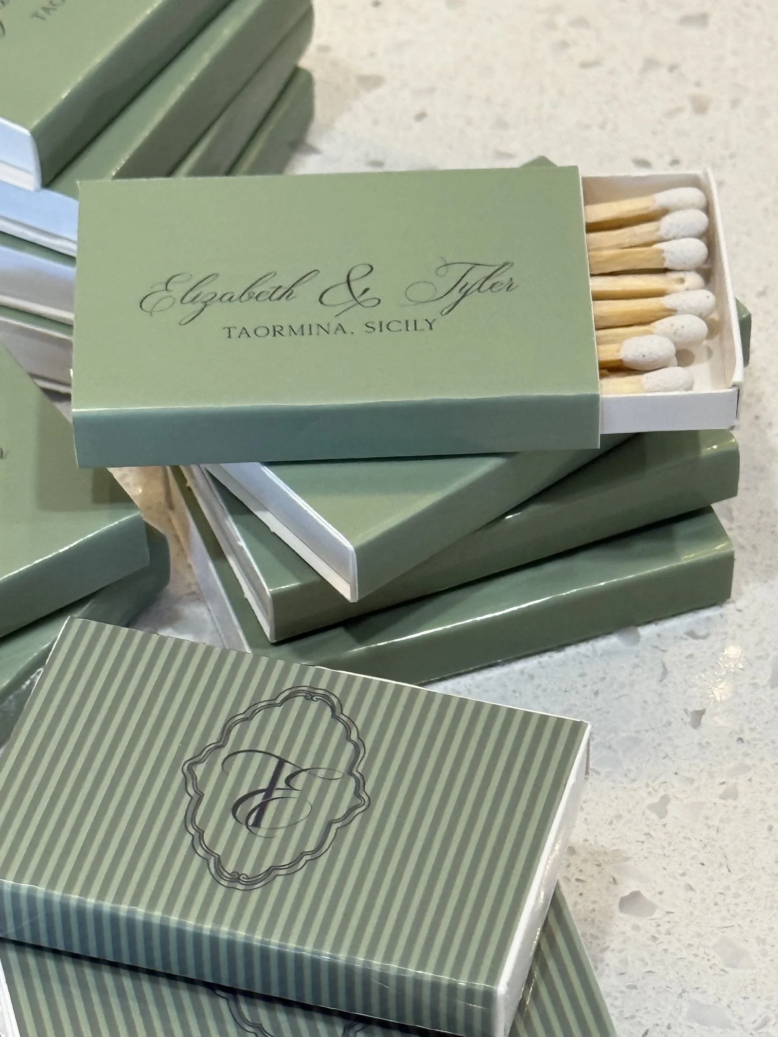 Box of matches with a light green cover and black decorative border, partially open, showing wooden matches with white tips inside.