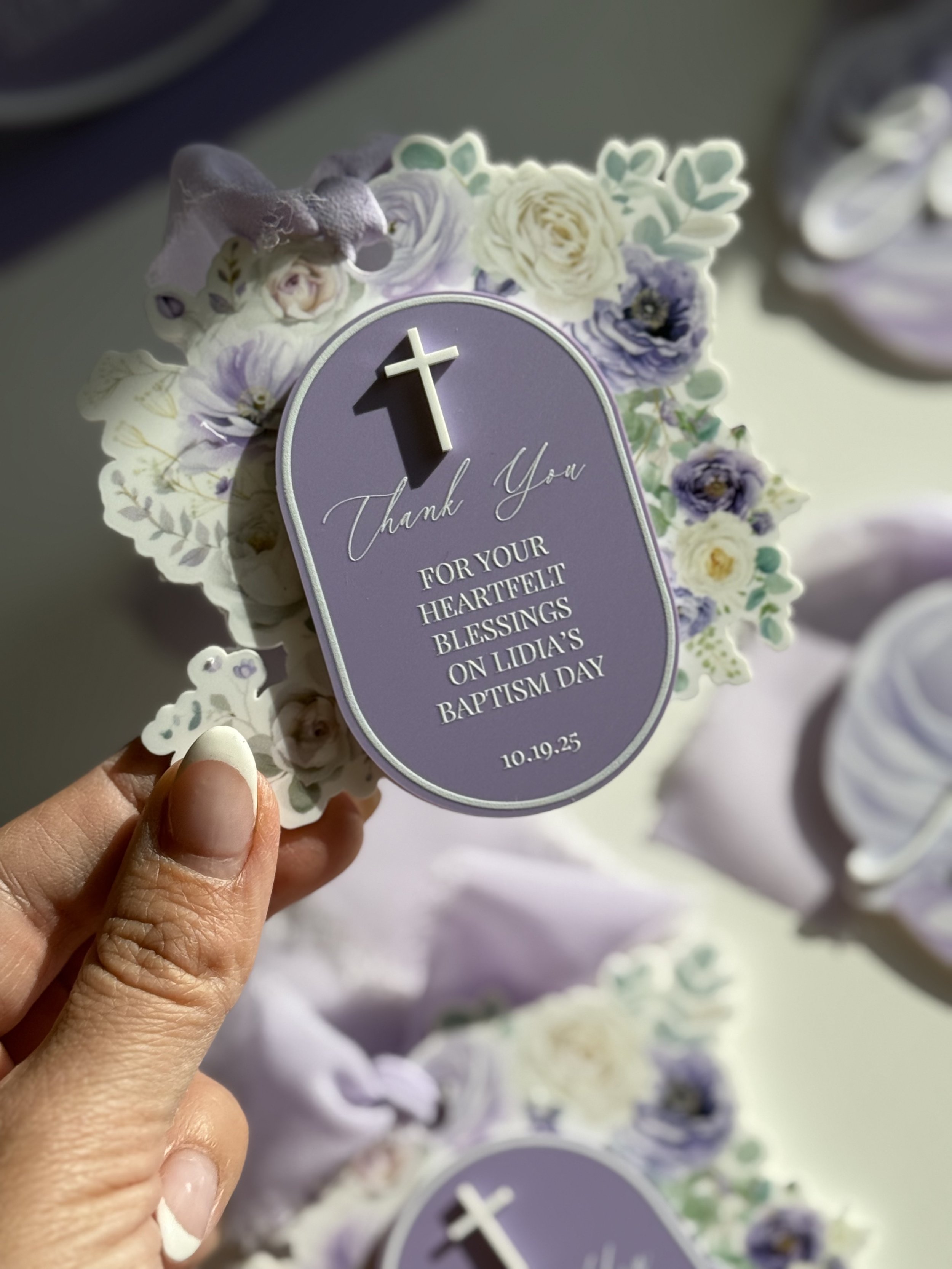 A hand holding a purple thank you card with a small cross on top, surrounded by floral decorations, for a baptism celebration.