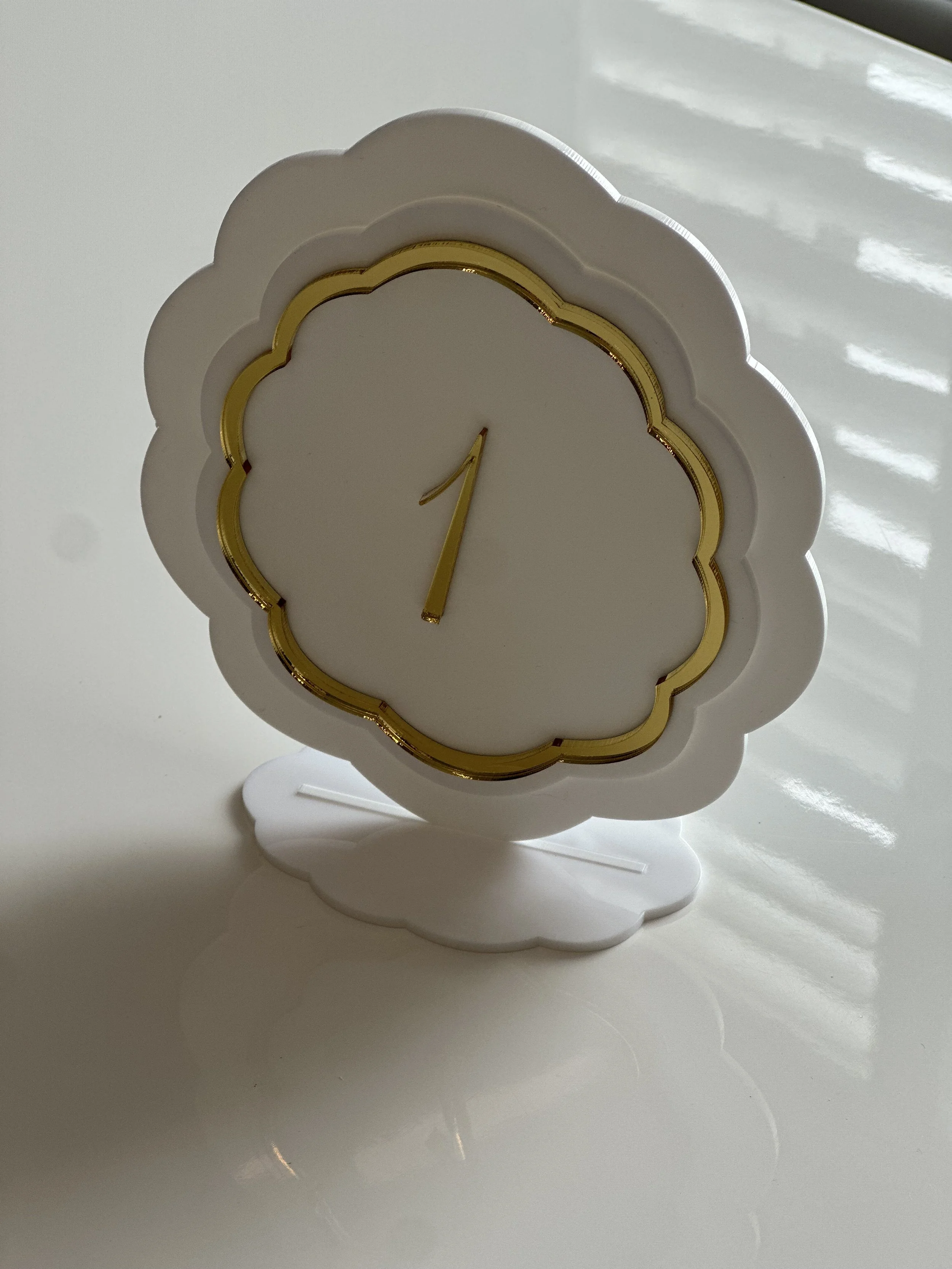 A decorative clock in the shape of a cloud with a white frame, gold detailing, and a minimalistic design, placed on a reflective white surface.