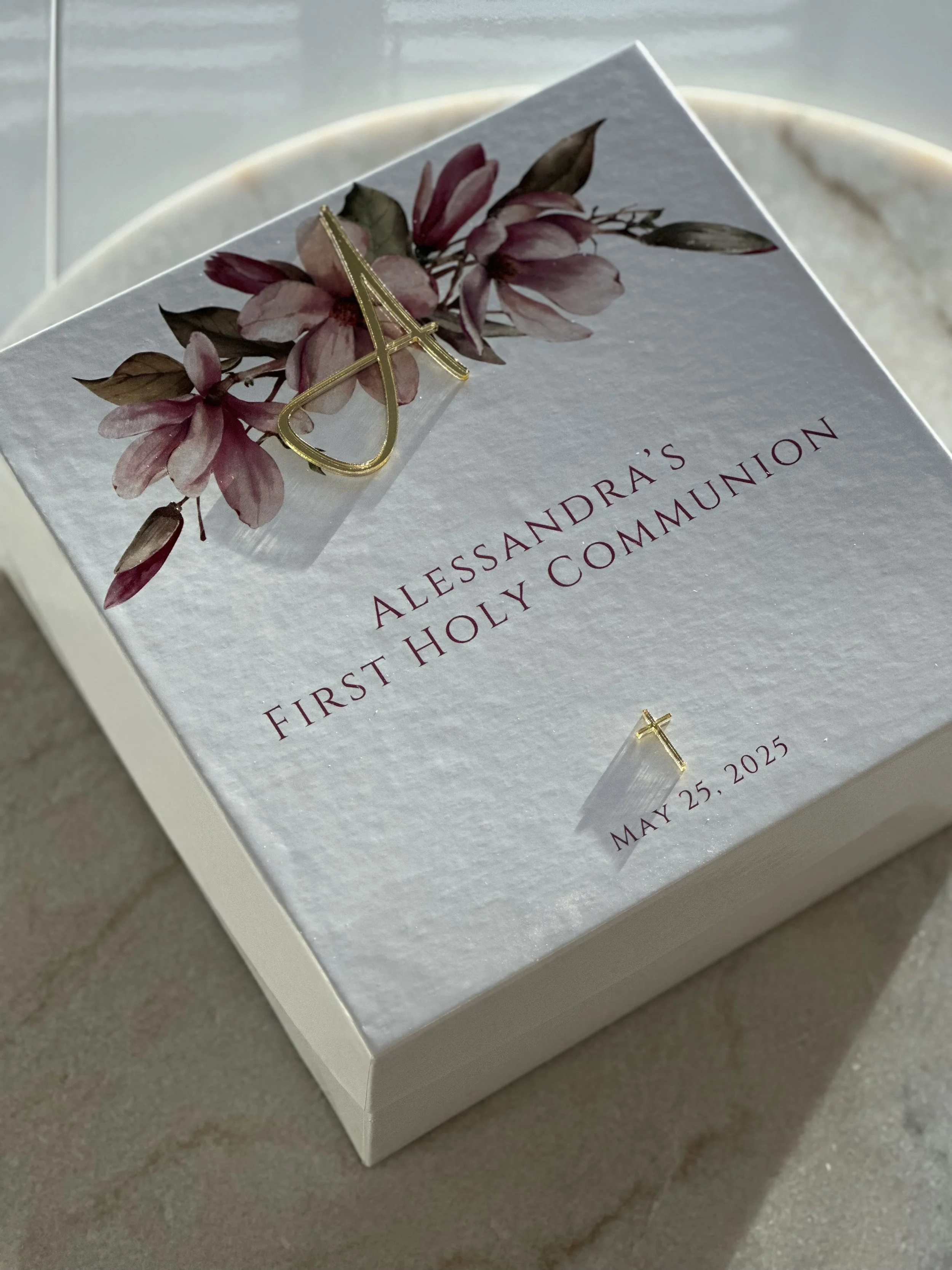 A white box with floral artwork and gold jewelry, including a cross pendant and initials, for a First Holy Communion celebration on May 25, 2025.