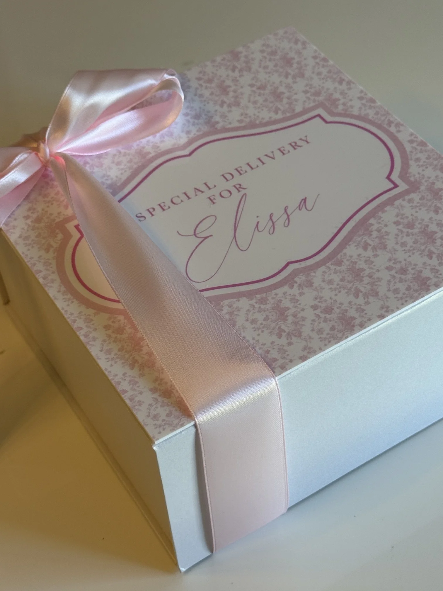 White gift box decorated with a light pink satin ribbon and a pink floral pattern on the lid, with a label reading 'Special Delivery for G Lisza' in pink and black.