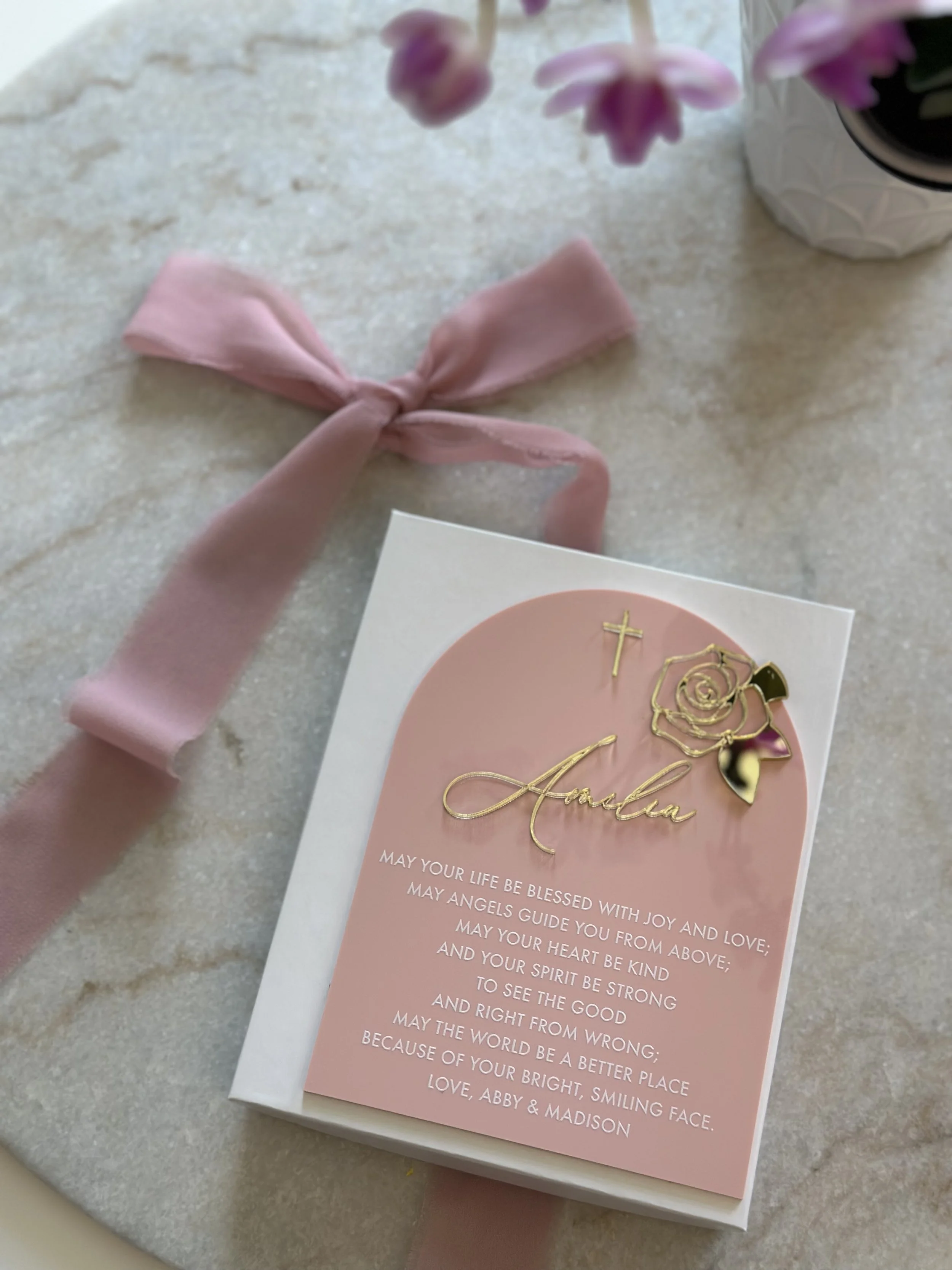 A greeting card with a pink background, decorative gold embellishments, and a gold cutout of a rose, cross, and the name 'Amala'. The card is placed on a white box, with a pink ribbon bow and a vase with pink flowers nearby.
