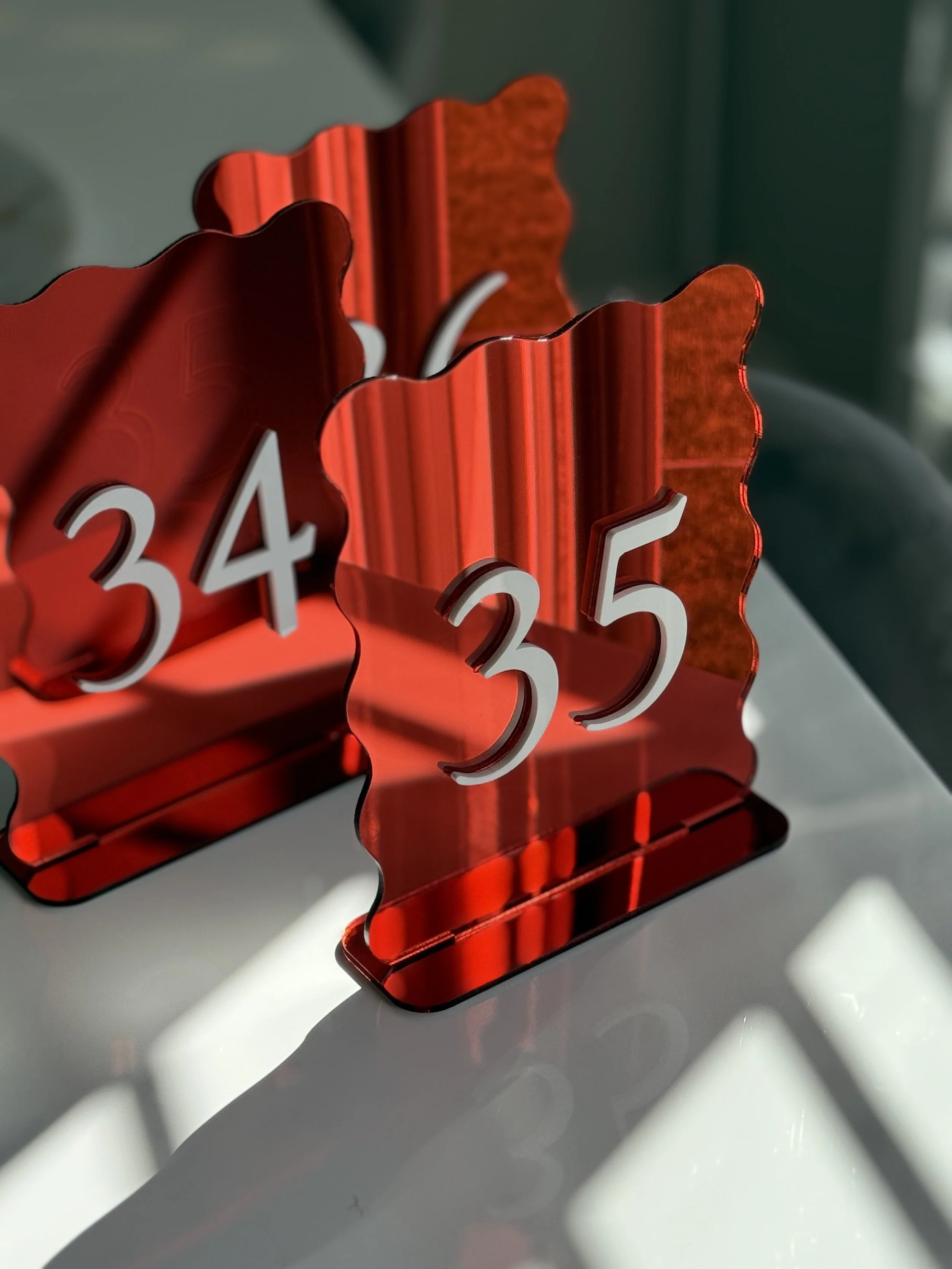 Red decorative table number holders with white numerals 34 and 35, reflecting on a white surface, with shadows cast on the surface.