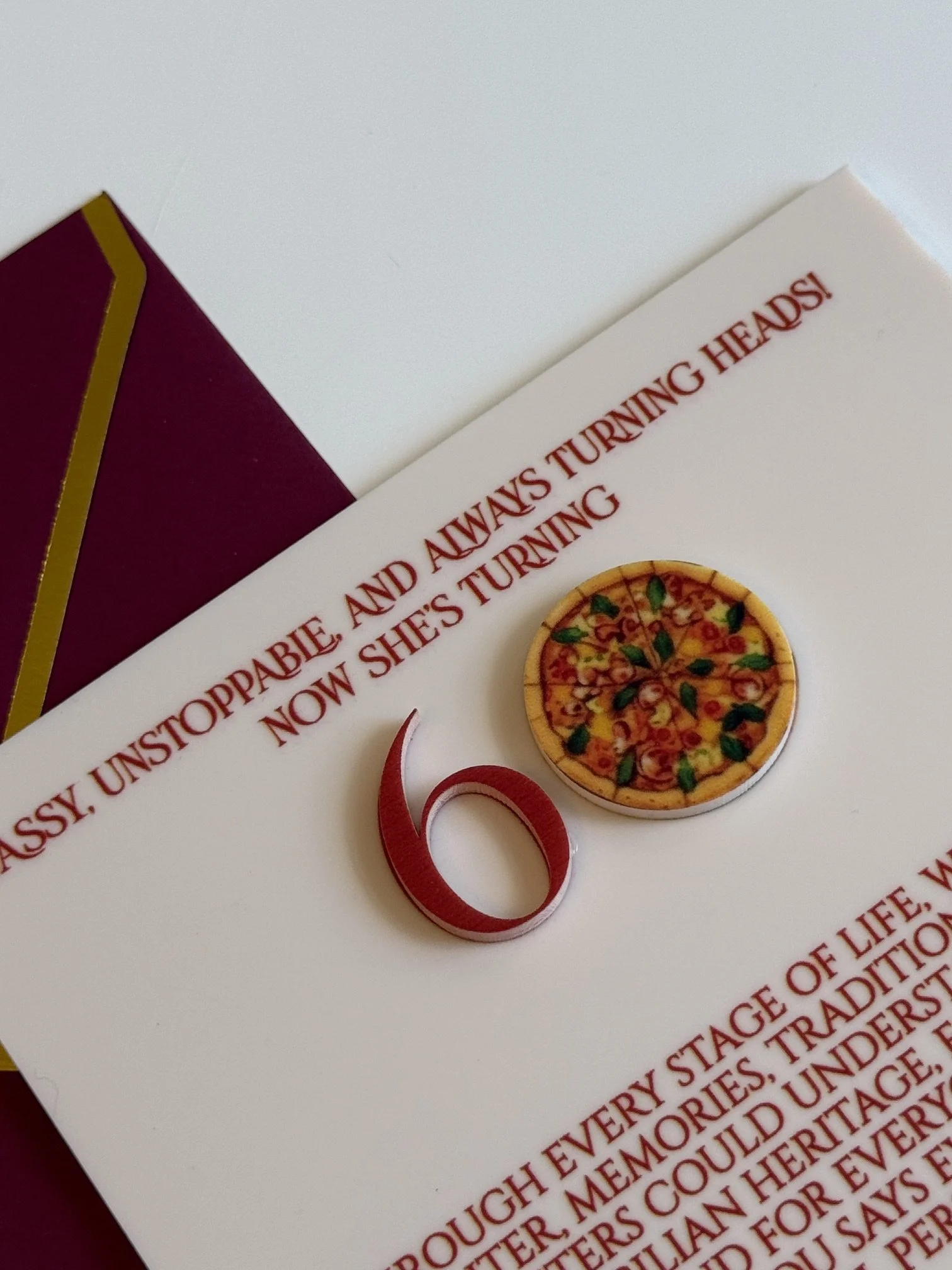 A white piece of paper with red text and a small circular image of a pizza. There is a red ribbon on the paper. Part of the maroon and gold border of a box is visible in the top left corner.