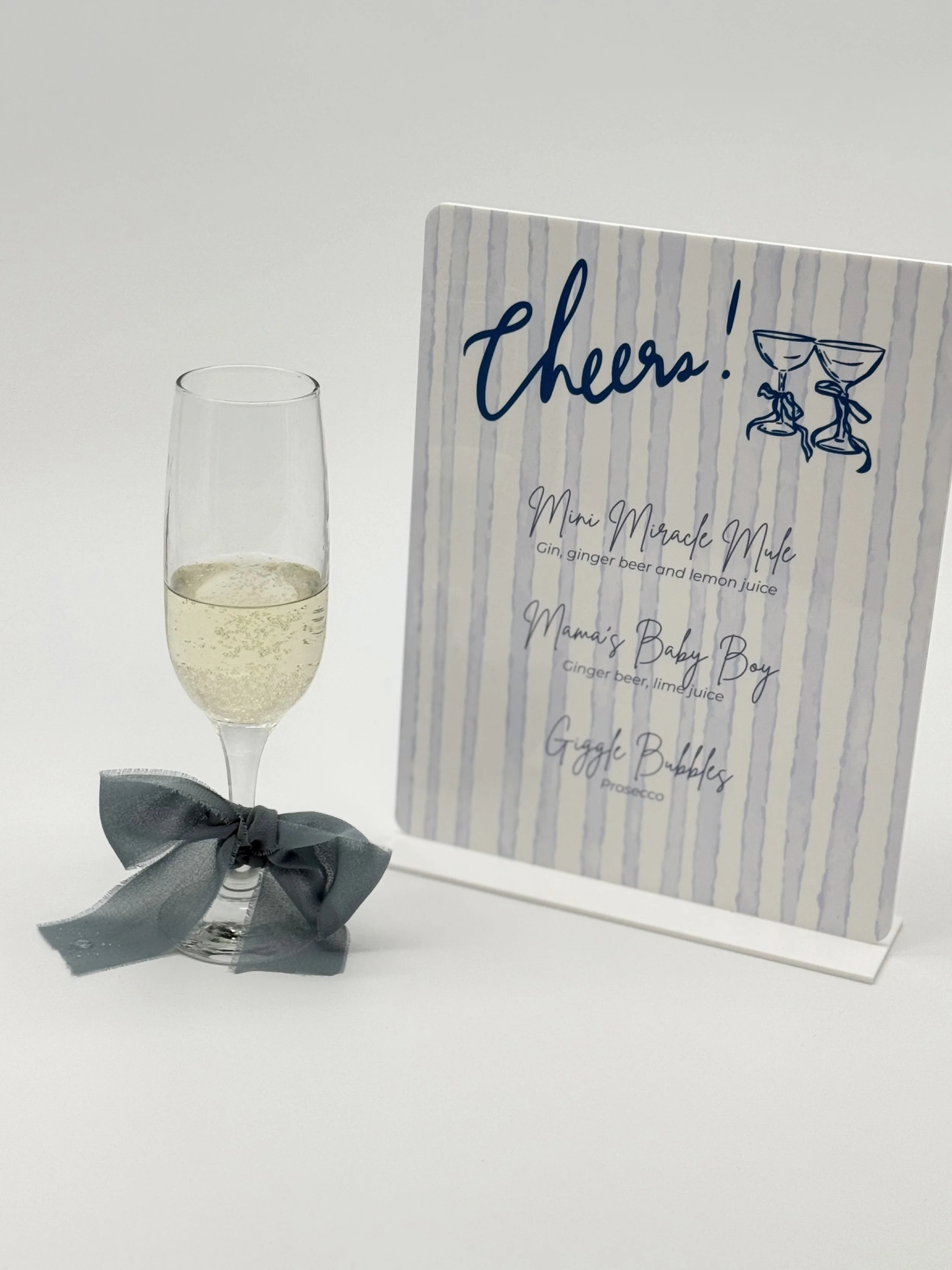 A glass of sparkling wine with a gray ribbon tied around the stem, placed beside a table sign with a striped background and handwritten-style text that reads 'Cheers!' and a drawing of two clinking glasses, listing three drink options.