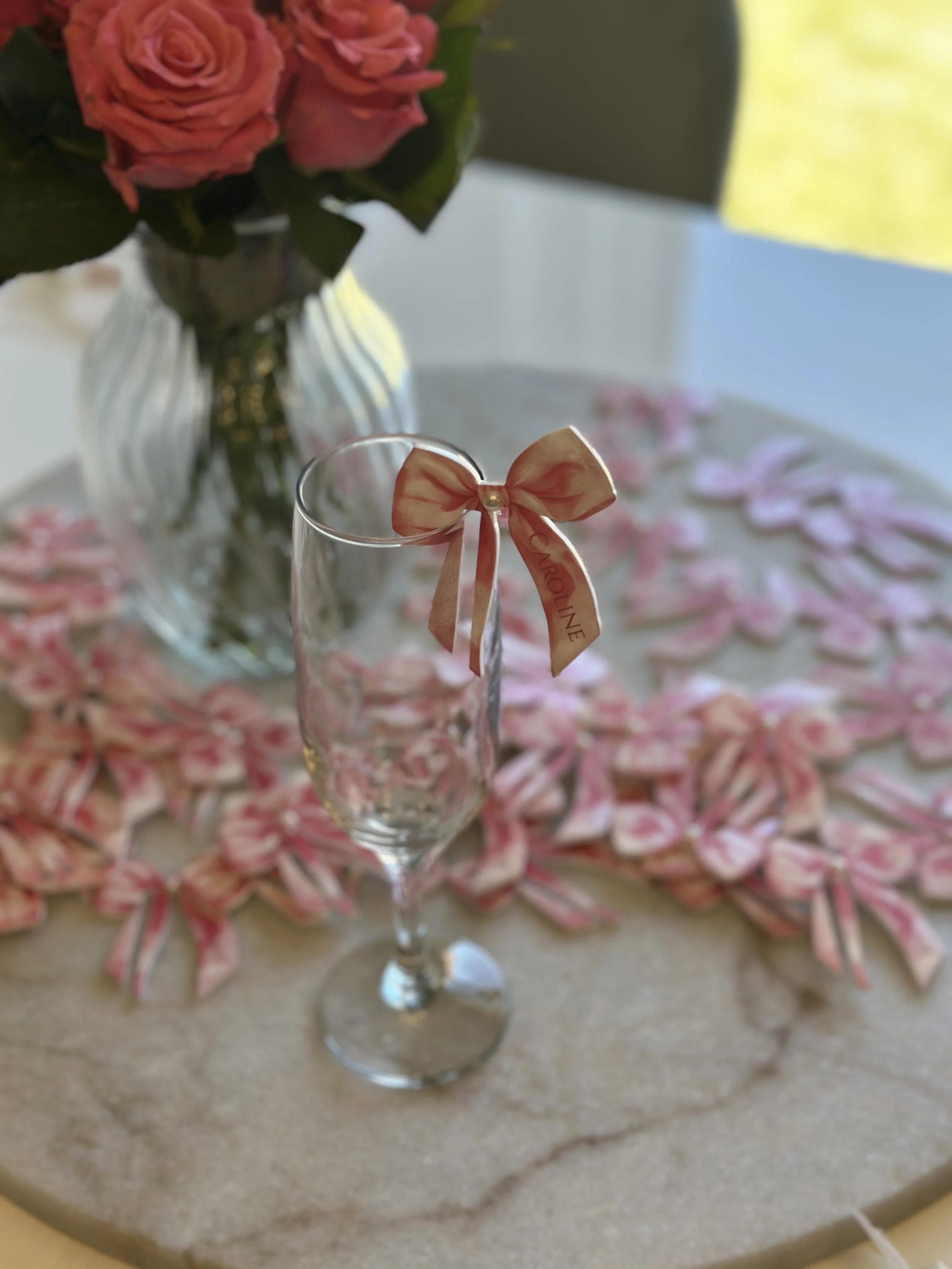 A champagne flute with a peach-colored ribbon tied in a bow around the rim, placed on a marble surface with pink and white wrapped candies scattered around. In the background, there is a glass vase with pink roses and green leaves.