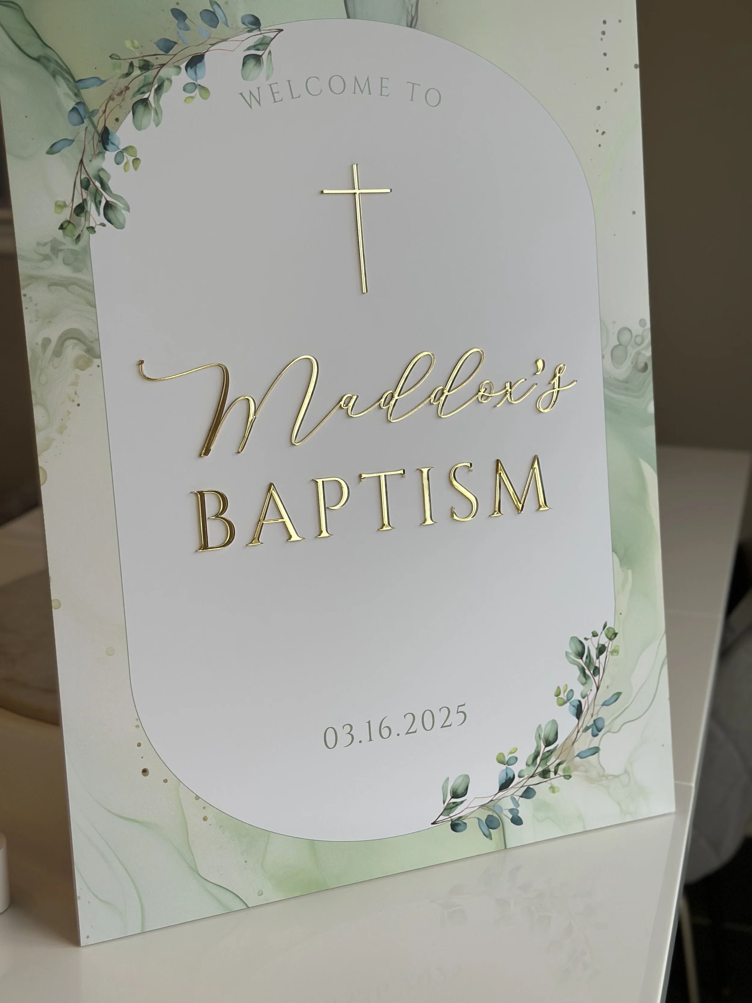Welcome sign for Maddox's baptism with a gold cross, date 03.16.2025, and green leaf decorations.