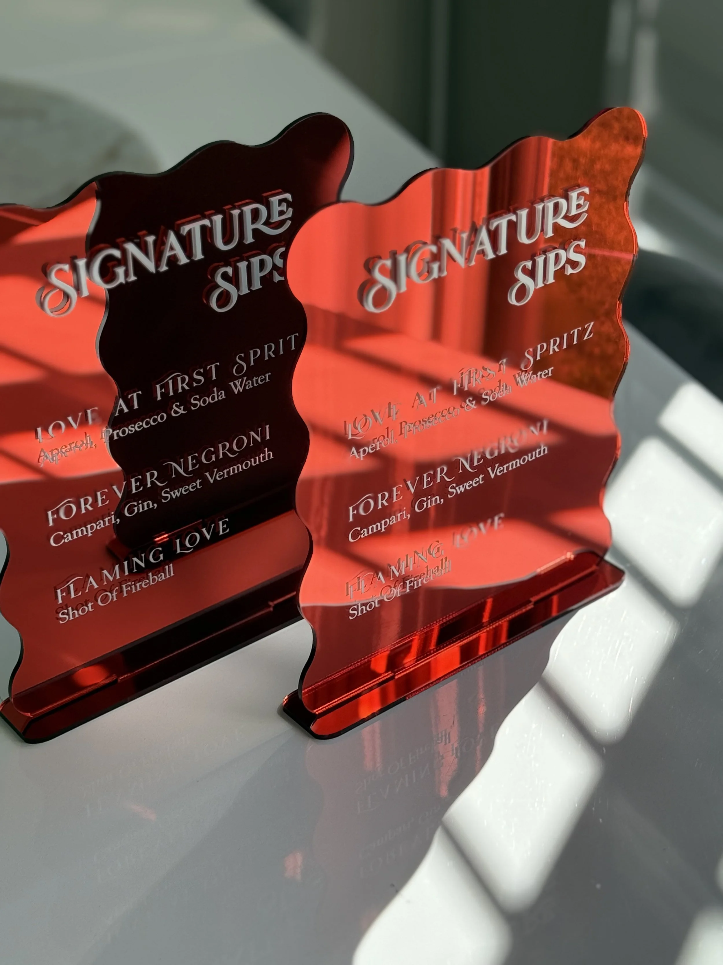Red acrylic napkin holders with the words 'Signature Sips' and various cocktail names, placed on a white surface near a window letting in natural light.