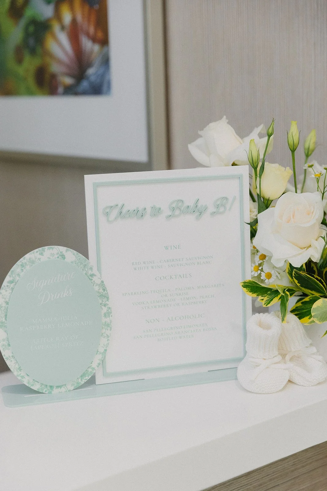 A table with a white sign that reads 'Cheers to Baby B!', a round sign with drink options, and a floral arrangement with white flowers and green leaves.
