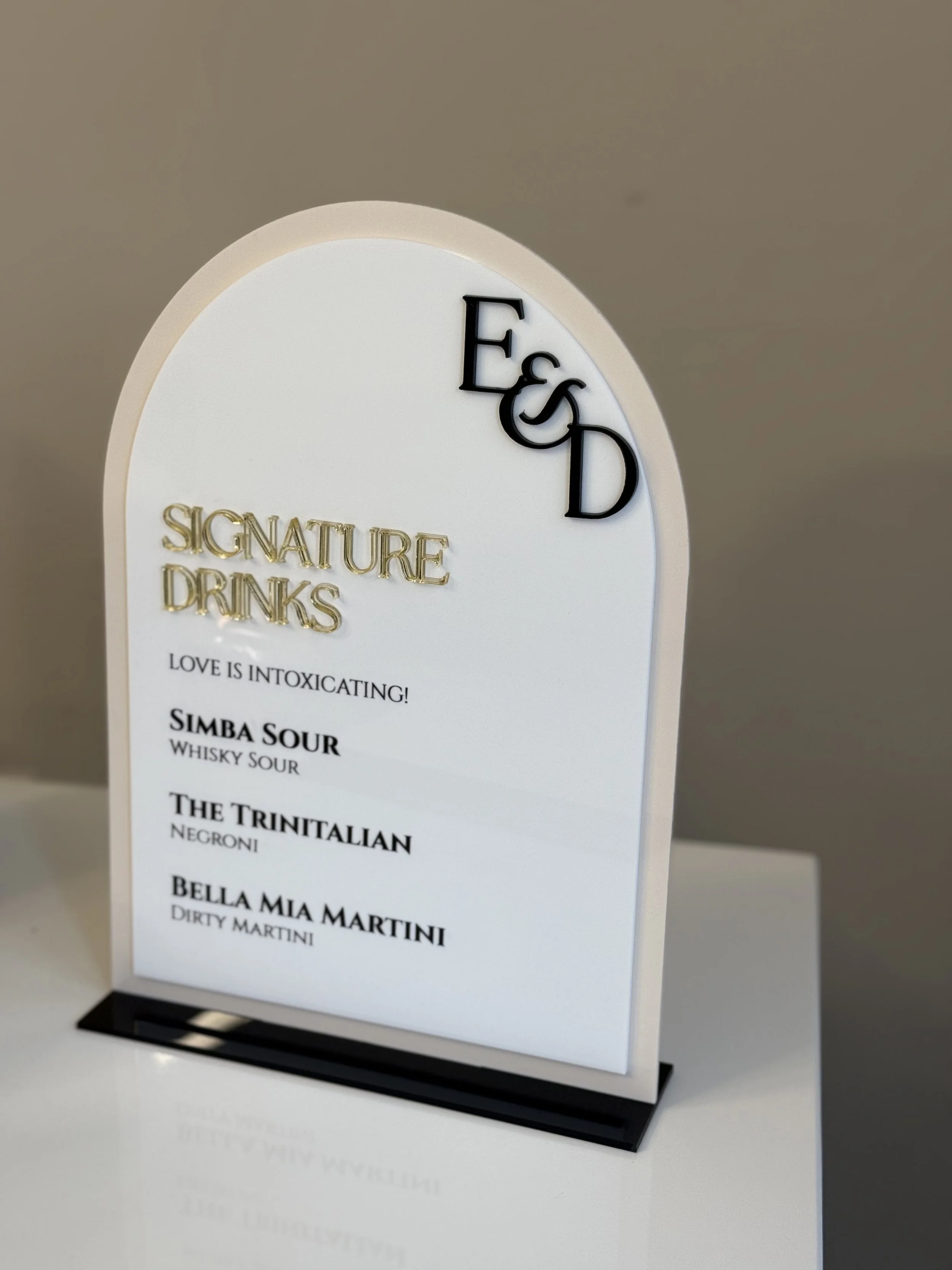 White signboard with black 3D letters 'E&S D' at the top right corner and gold letters 'Signature Drinks', listing three drinks: 'Simba Sour', 'The Trinitalian', and 'Bella Mia Martini' with their descriptions.