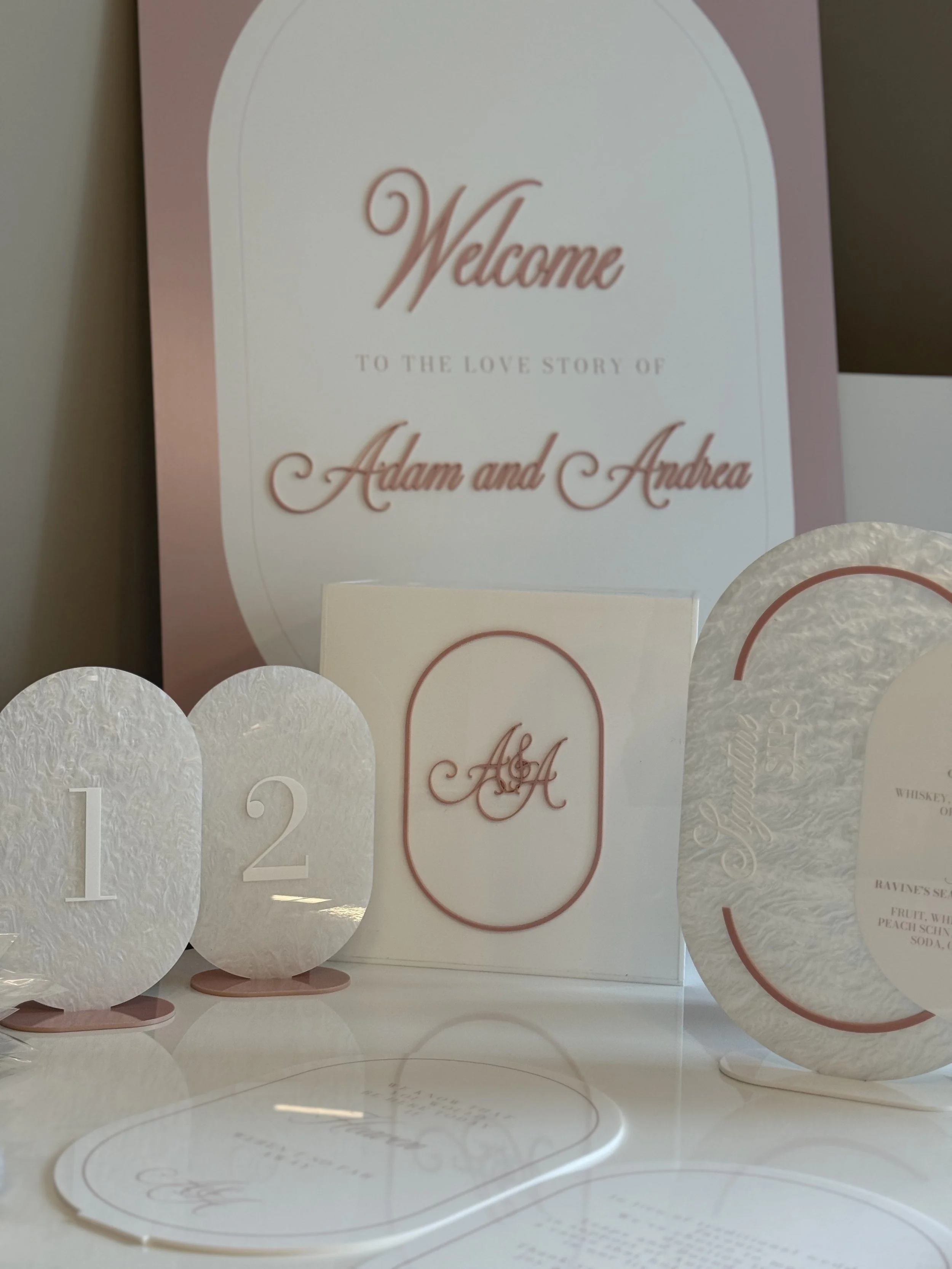 Wedding decor including a large welcome sign for Adam and Andrea, table numbers 1 and 2, and an initials sign 'A & A' in pink and white colors.