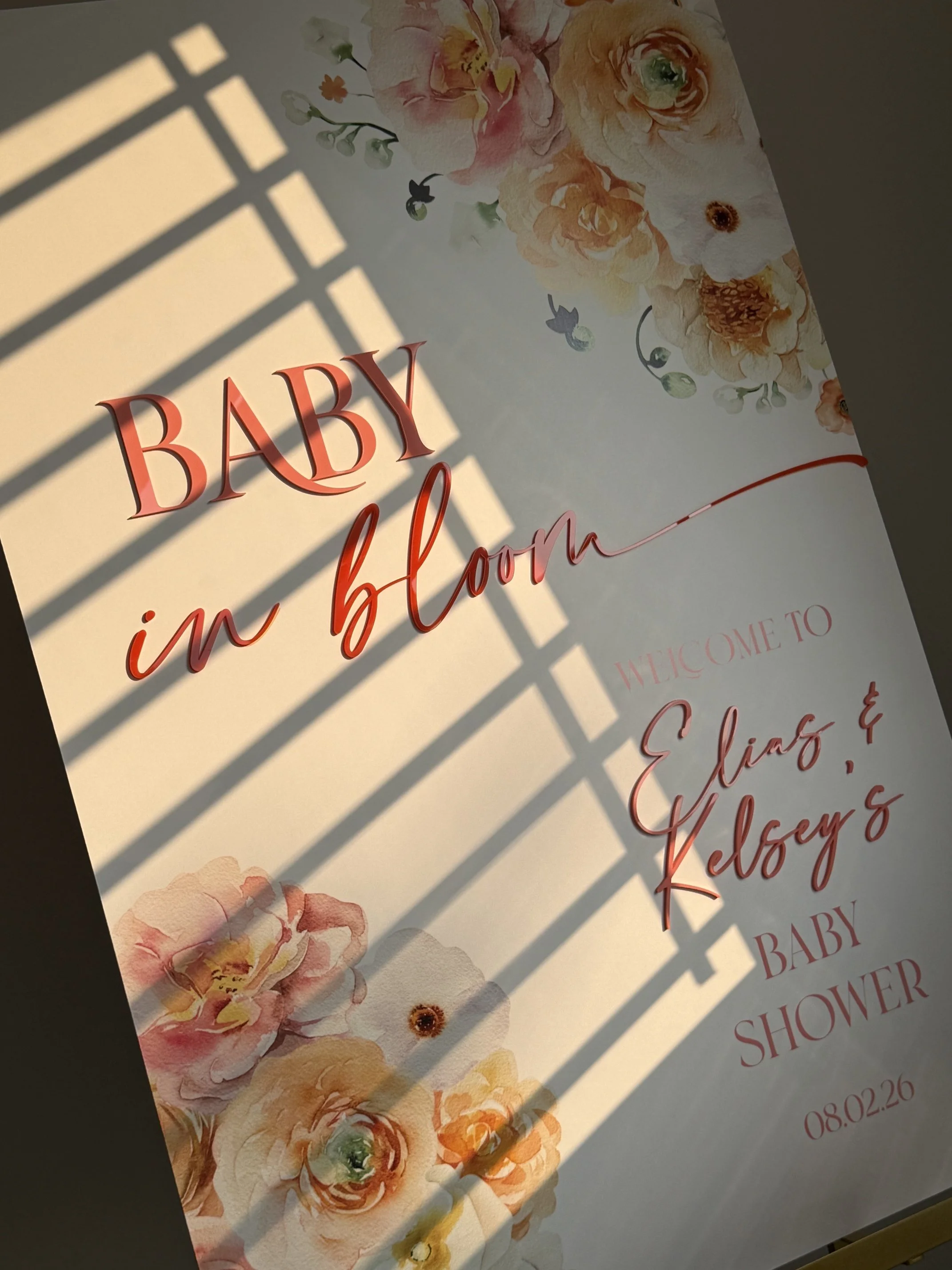 A floral wedding shower invitation with a floral design at the top and bottom, and text in red and pink fonts. The visible text reads 'BABY in bloom,' and includes the date 08.02.26.