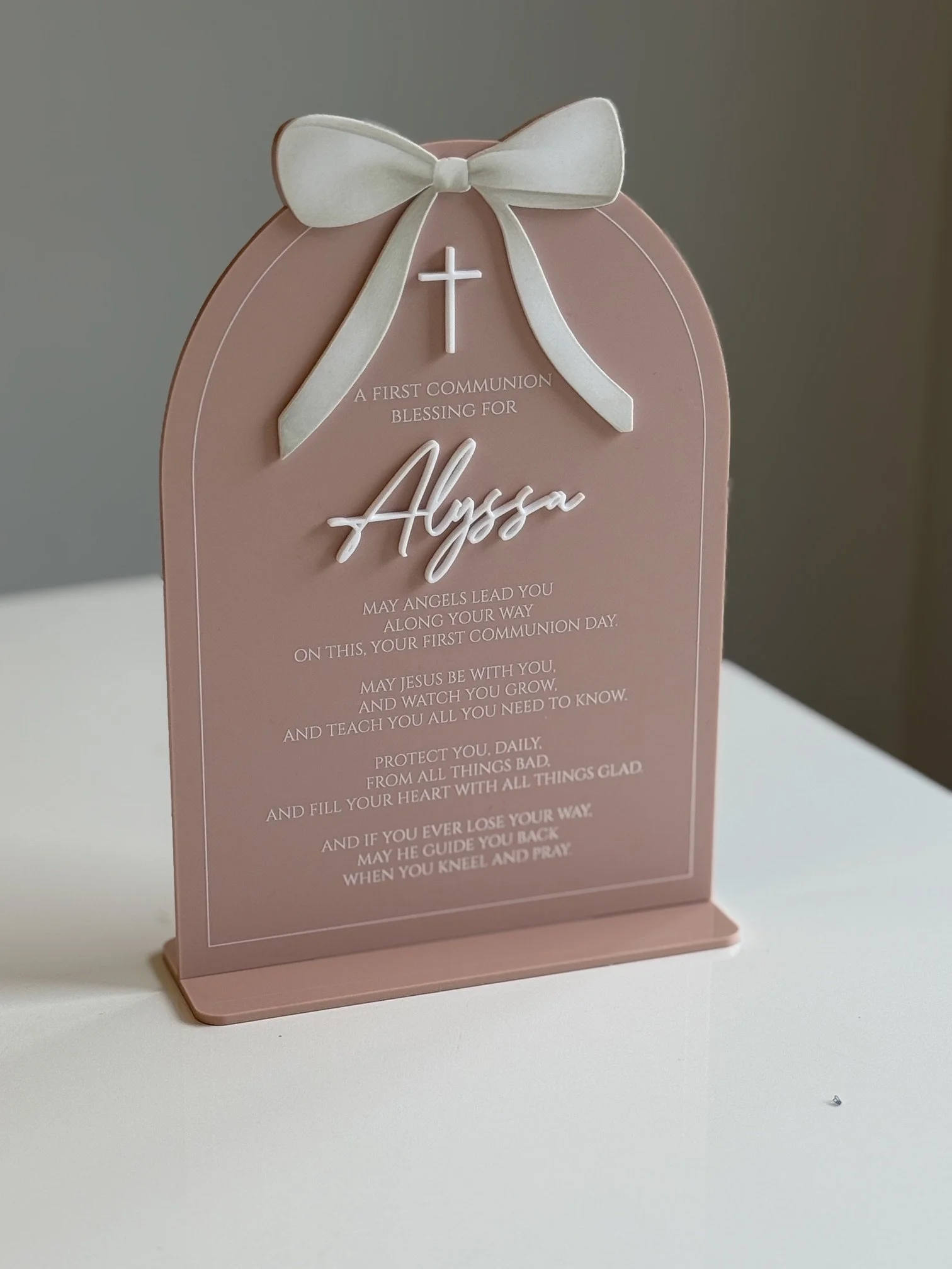 A pink decorative stand with a white bow on top displaying a prayer for a child's first communion. The prayer mentions blessings, guidance, and protection from Jesus and angels.