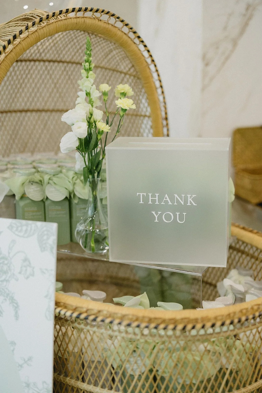 A thank you card on a glass table surrounded by white flower petals, a glass vase with white and yellow flowers, and jars with green labels in the background, all on a wicker chair.