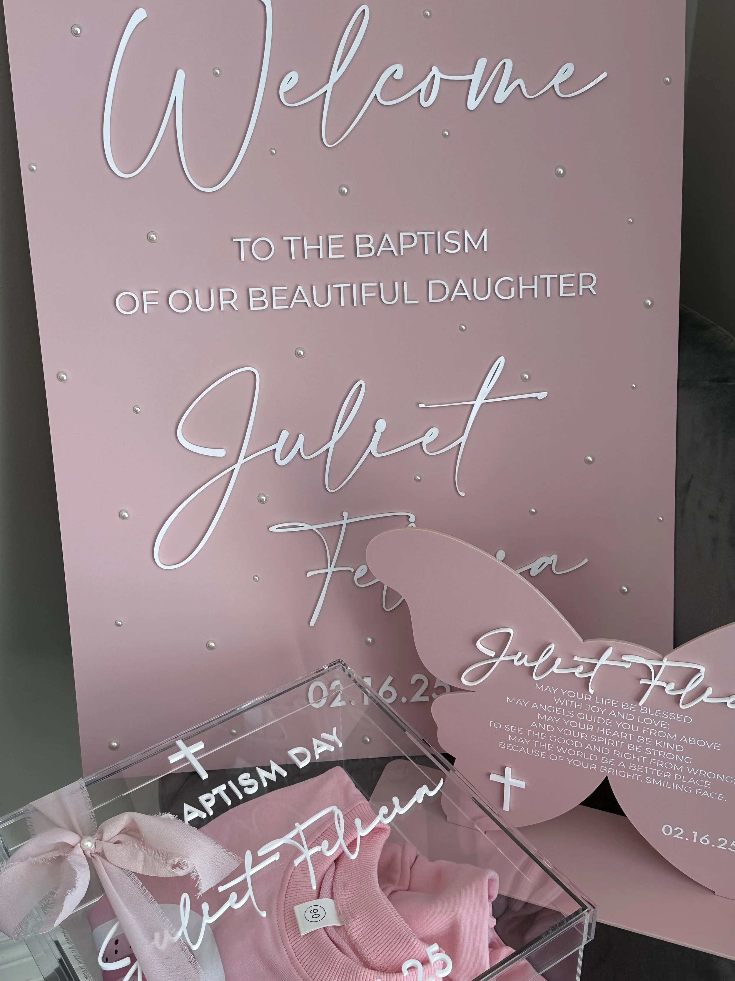 Pink welcome sign for a daughter's baptism, decorated with pearls and script fonts, with a pink baptism gown and a butterfly-themed card in front.